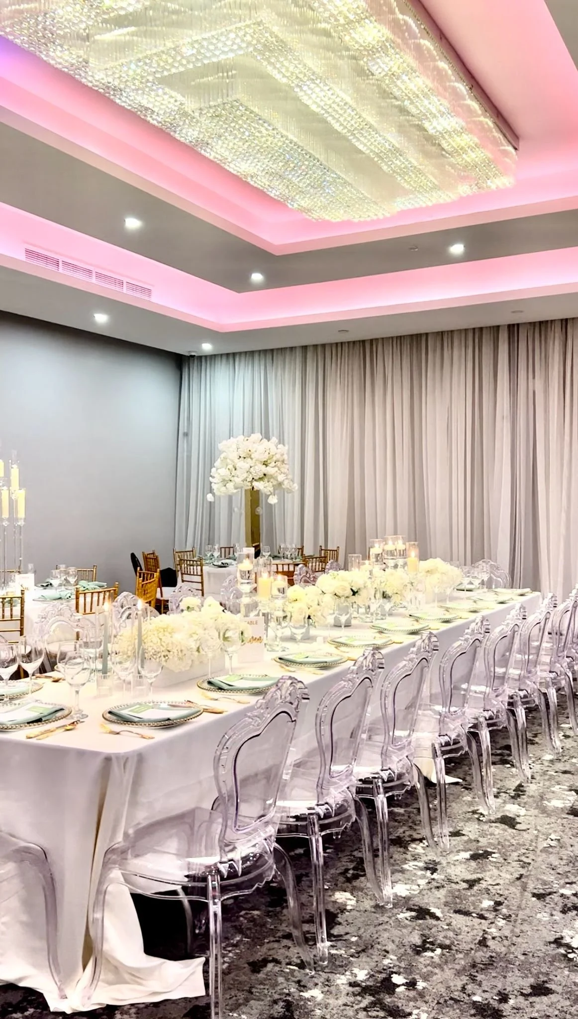Elegant banquet hall with a long table decorated with white flowers, candles, and fine tableware, surrounded by transparent chairs, with a large floral centerpiece and draped curtains in the background.