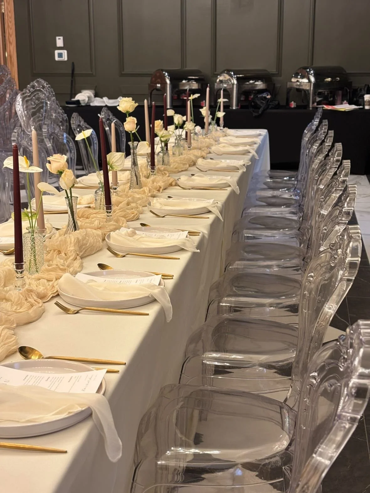 Elegant long dining table set for a formal event with white tablecloth, gold utensils, white napkins, small floral centerpieces, and tall candles, surrounded by transparent chairs in a decorated room.