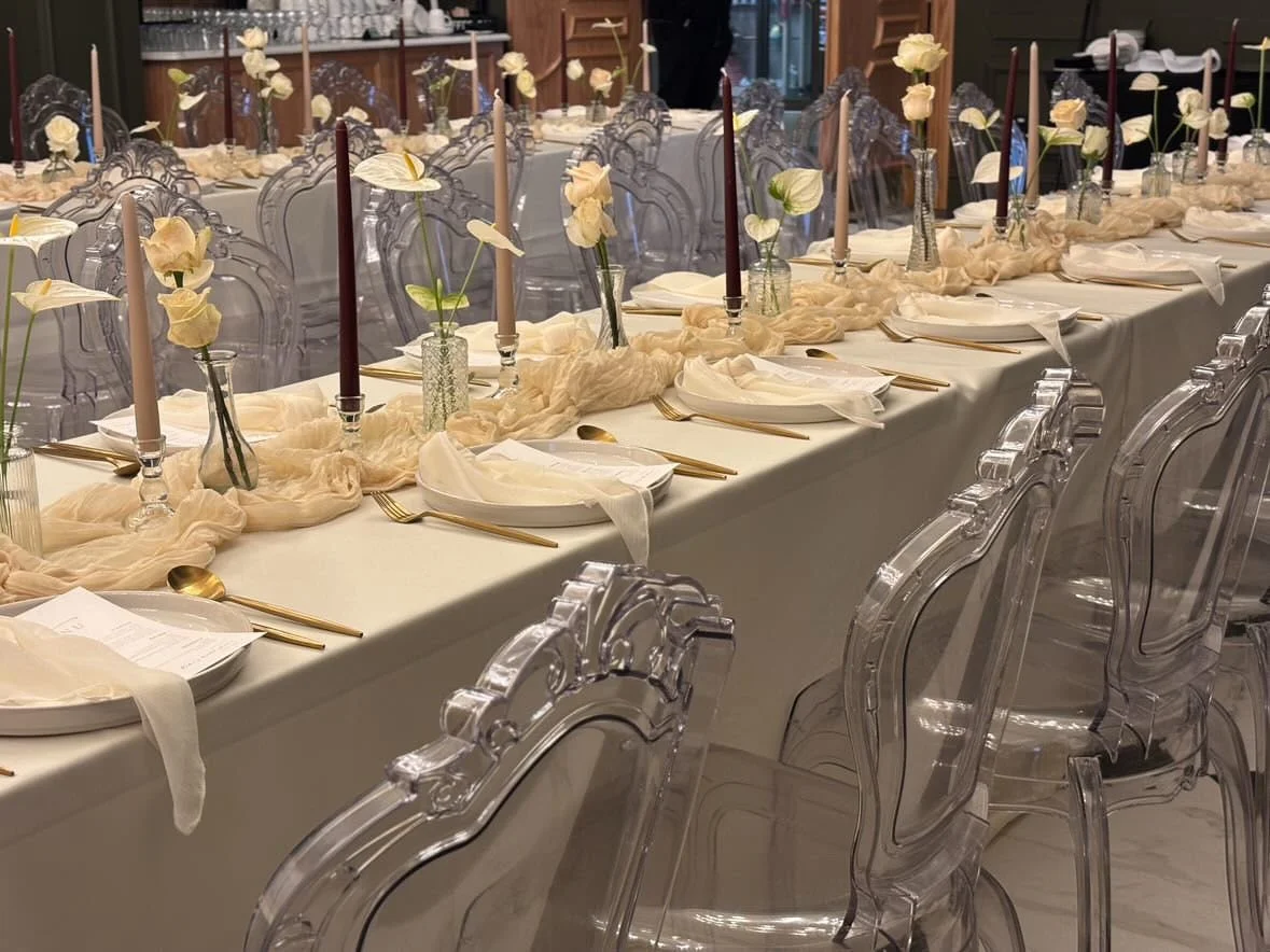 Elegant banquet table decorated with white flowers, beige cloth, gold cutlery, purple candles, and soft fabric draped along the center, transparent chairs lining the sides.