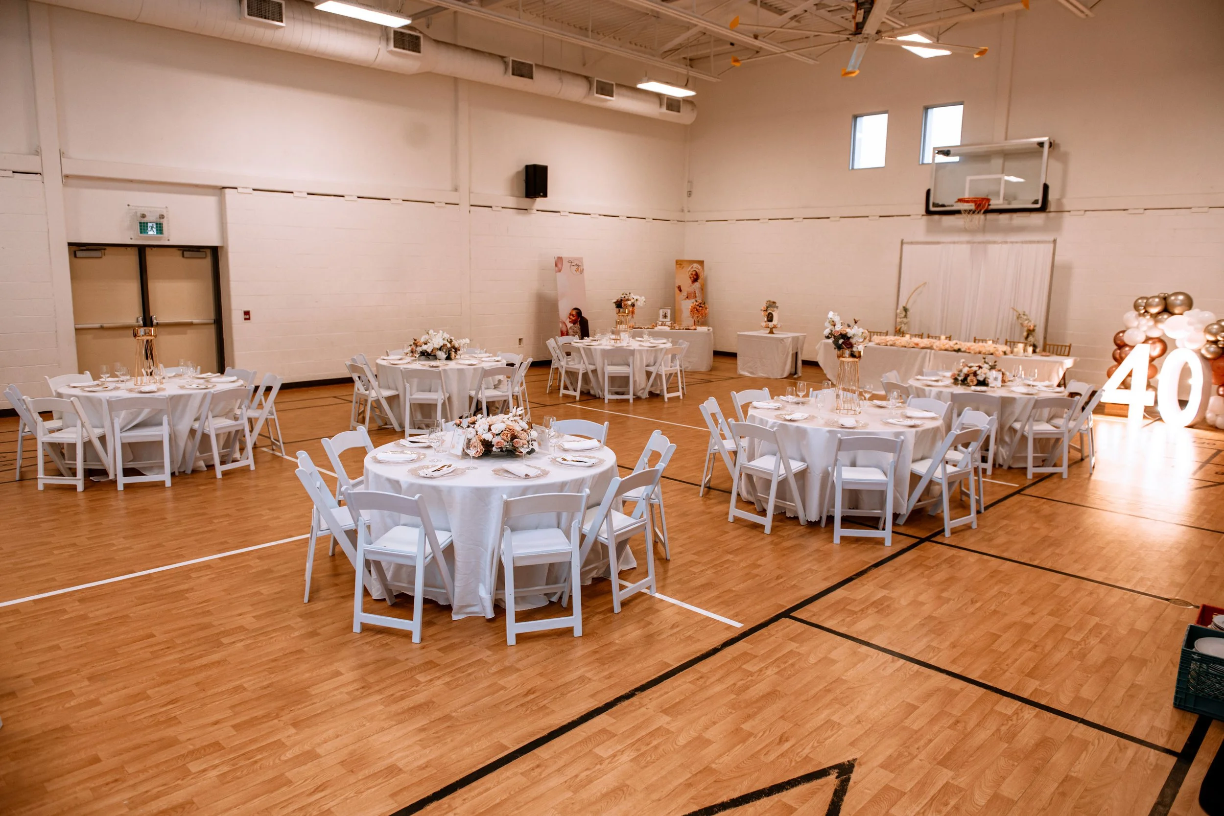 Decorated banquet tables with white tablecloths, floral centerpieces, and place settings in a gymnasium with wooden floor, basketball hoop, and large illuminated number 40.