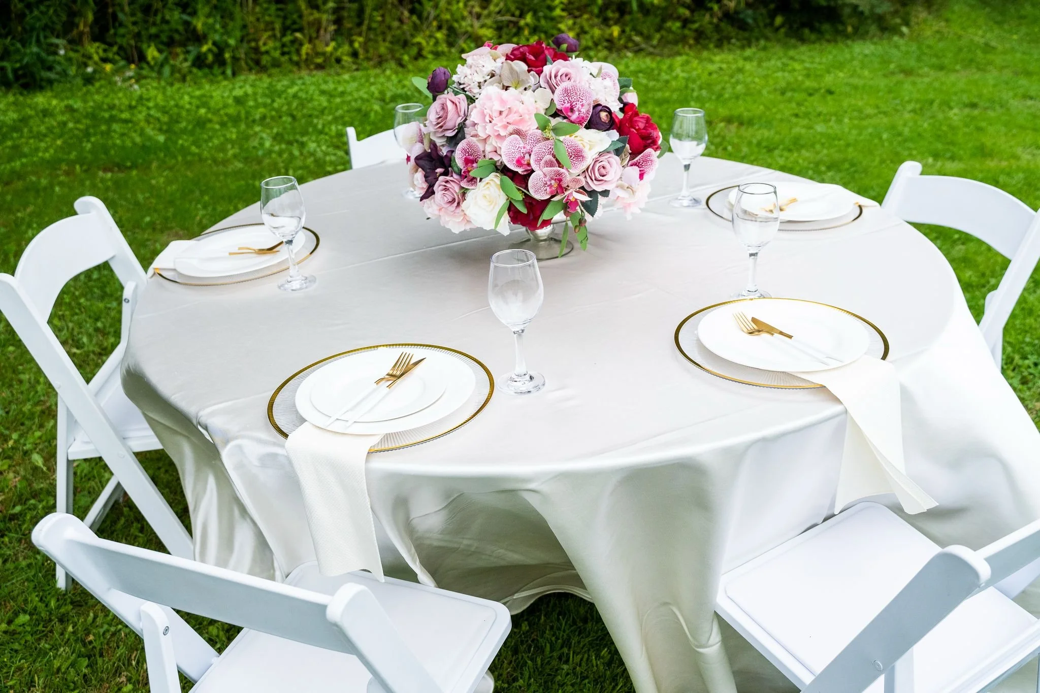 Round table set for outdoor event with a cream tablecloth, four white chairs, a large pink and white floral centerpiece, white plates with gold-rimmed edges, white napkins, and glasses of water, on a grassy lawn.