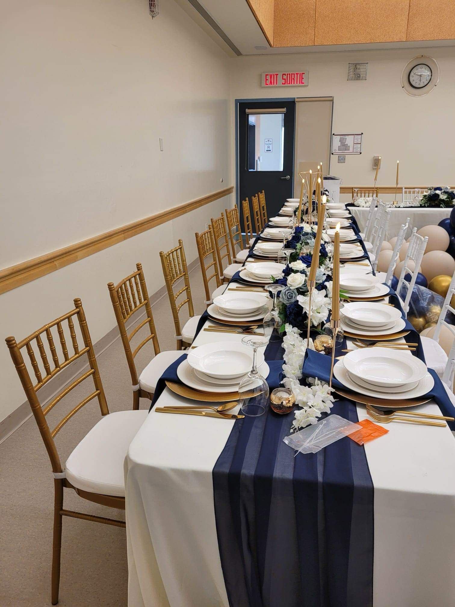 Long dining table set for a celebration with white plates, gold cutlery, navy blue and white floral centerpiece, tall gold candles, and wine glasses, in a decorated event space.