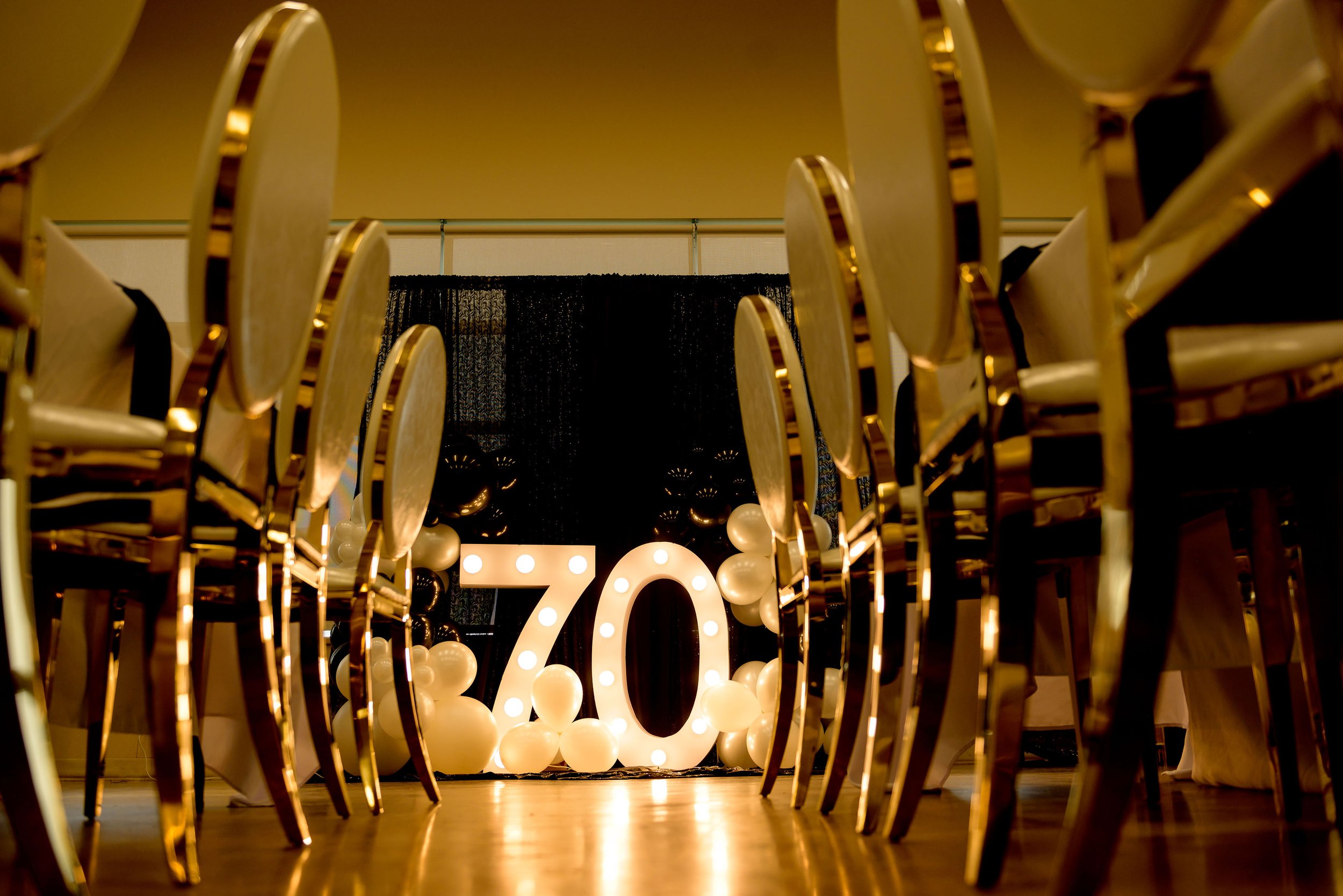 A banquet hall decorated for a 70th birthday celebration with large illuminated number 70 and white balloons, with chairs arranged around tables in the foreground.
