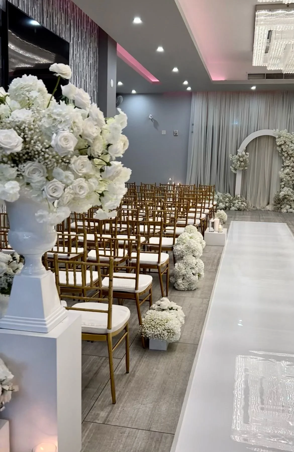 Elegant indoor wedding ceremony setup with gold chairs, white floral arrangements, and a white aisle runner facing an floral arch backdrop.