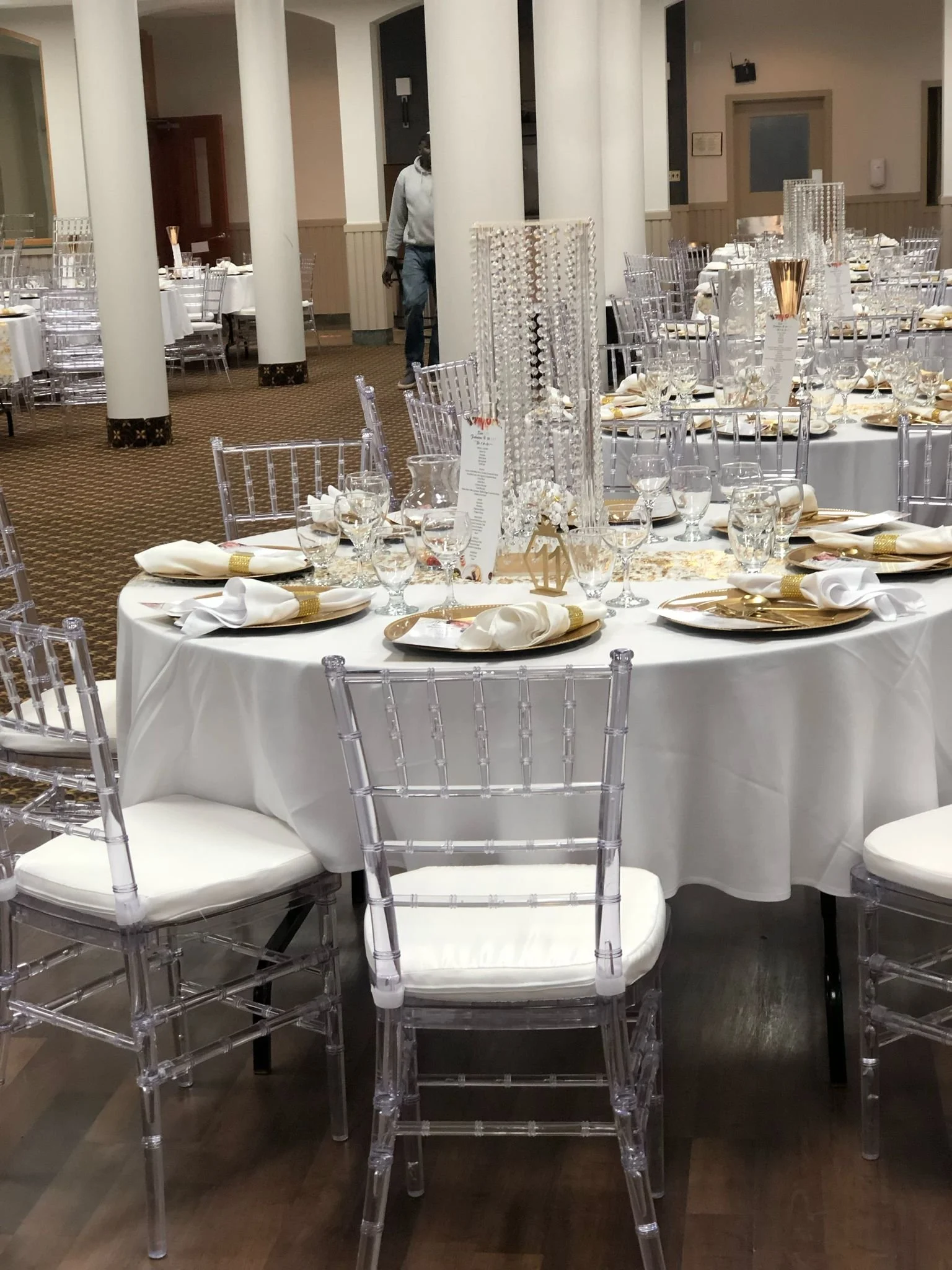 Elegant banquet table set for a wedding or formal event with white tablecloths, gold-rimmed plates, white napkins, multiple glasses, and decorative centerpieces in a large decorated hall.