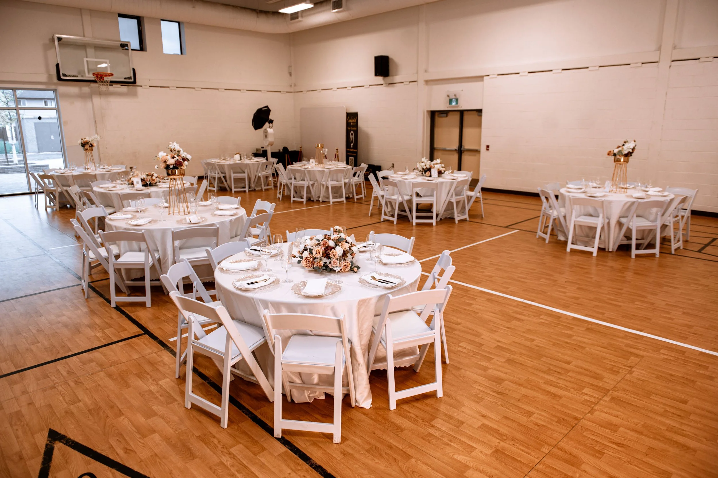 A banquet hall decorated for an event with round tables covered in white tablecloths and floral centerpieces, set with plates, glasses, and silverware. The room has a sports hall background with wooden flooring and white walls.