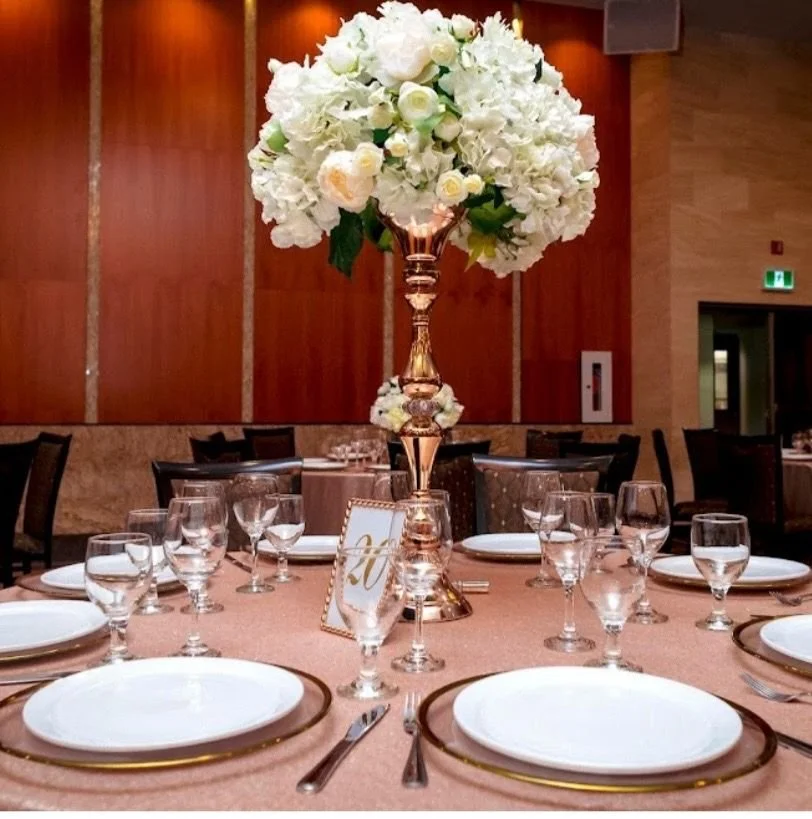 Elegant round banquet table set with white plates, water glasses, and silverware, featuring a tall floral centerpiece with white flowers in a gold vase, in a formal event hall with warm lighting and wooden panel walls.