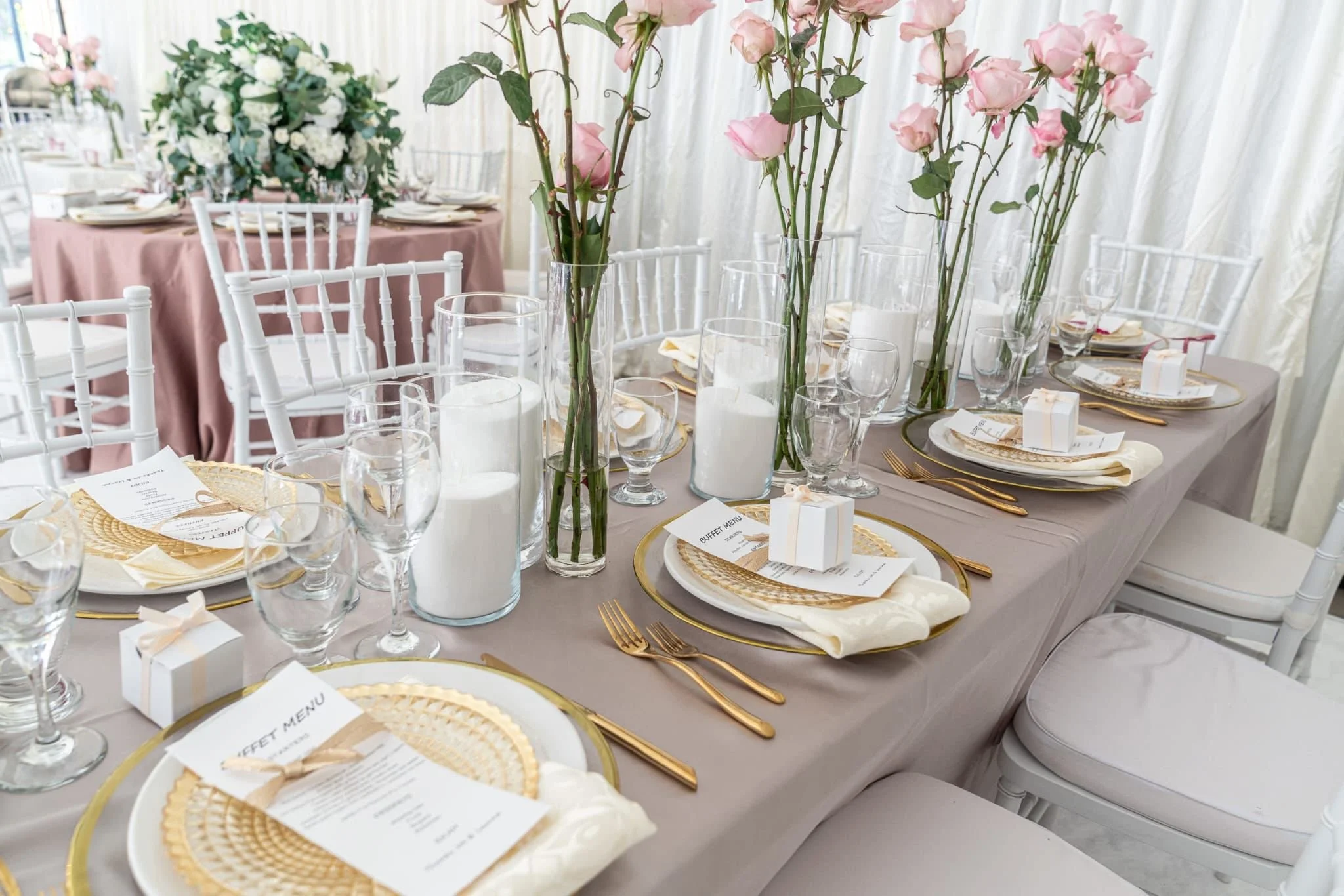 Elegant dining table set with pink and white floral centerpieces, candles, gold-rimmed plates, gold utensils, and white chairs, prepared for a special event or wedding reception.