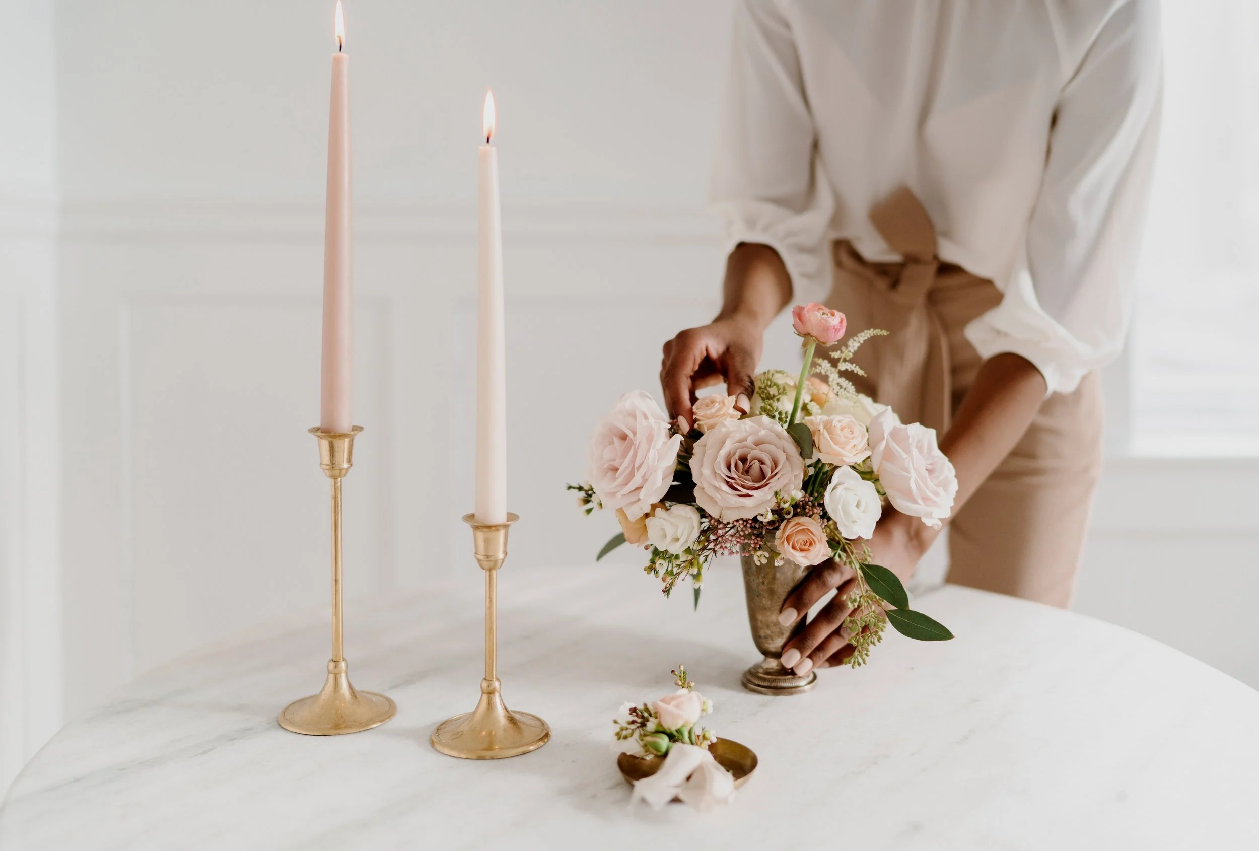 Person arranging a bouquet of pink and white flowers on a white table with two lit pink candles in gold candle holders nearby.