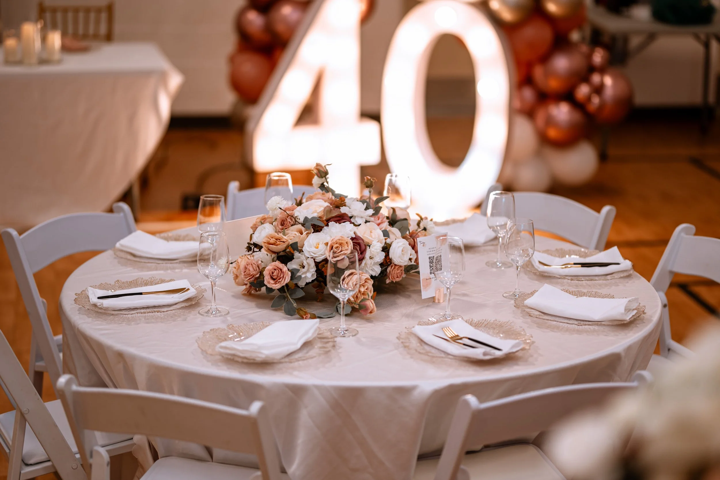 Round table decorated with a floral centerpiece, white plates, napkins, gold utensils, and wine glasses, set for a celebration. Large illuminated '40' sign with balloons is in the background.
