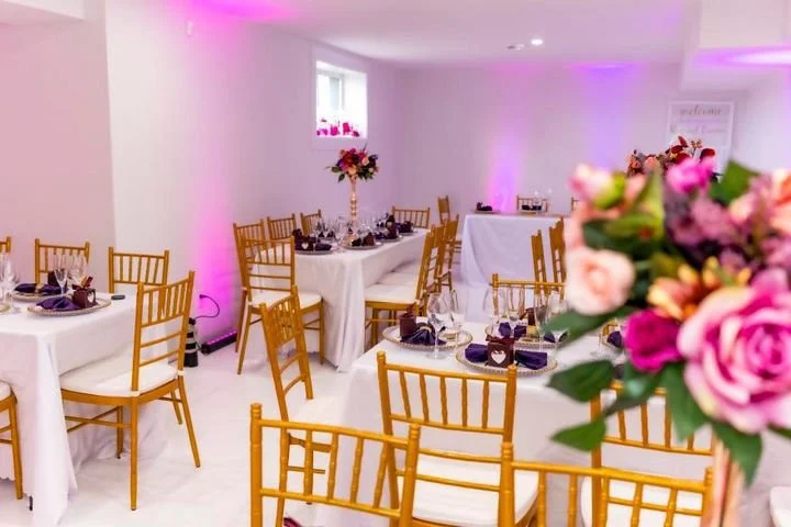 Wedding reception setup with round tables covered in white tablecloths, gold chairs, purple napkins, and floral centerpieces in a bright, well-lit room.