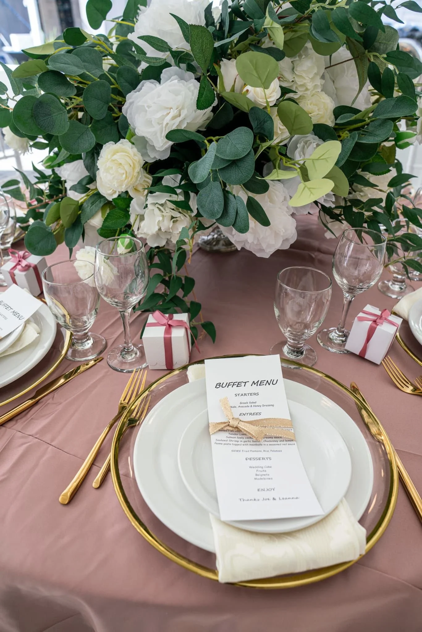 Elegant table setting with a large floral centerpiece of white flowers and greenery, gold-rimmed plates, gold cutlery, clear wine glasses, and a small white gift box with a pink ribbon, along with a white menu card on a pink tablecloth.