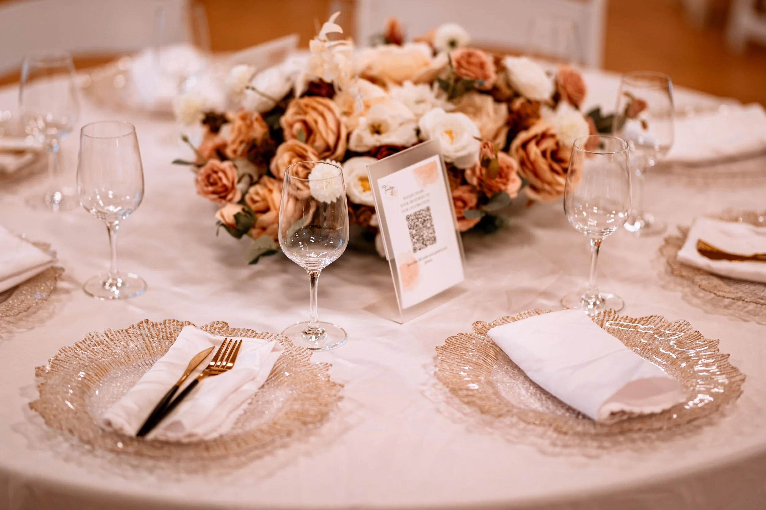 Elegant table setting with glassware, white napkins, gold utensils, and a large floral centerpiece with roses and greenery at a formal event.