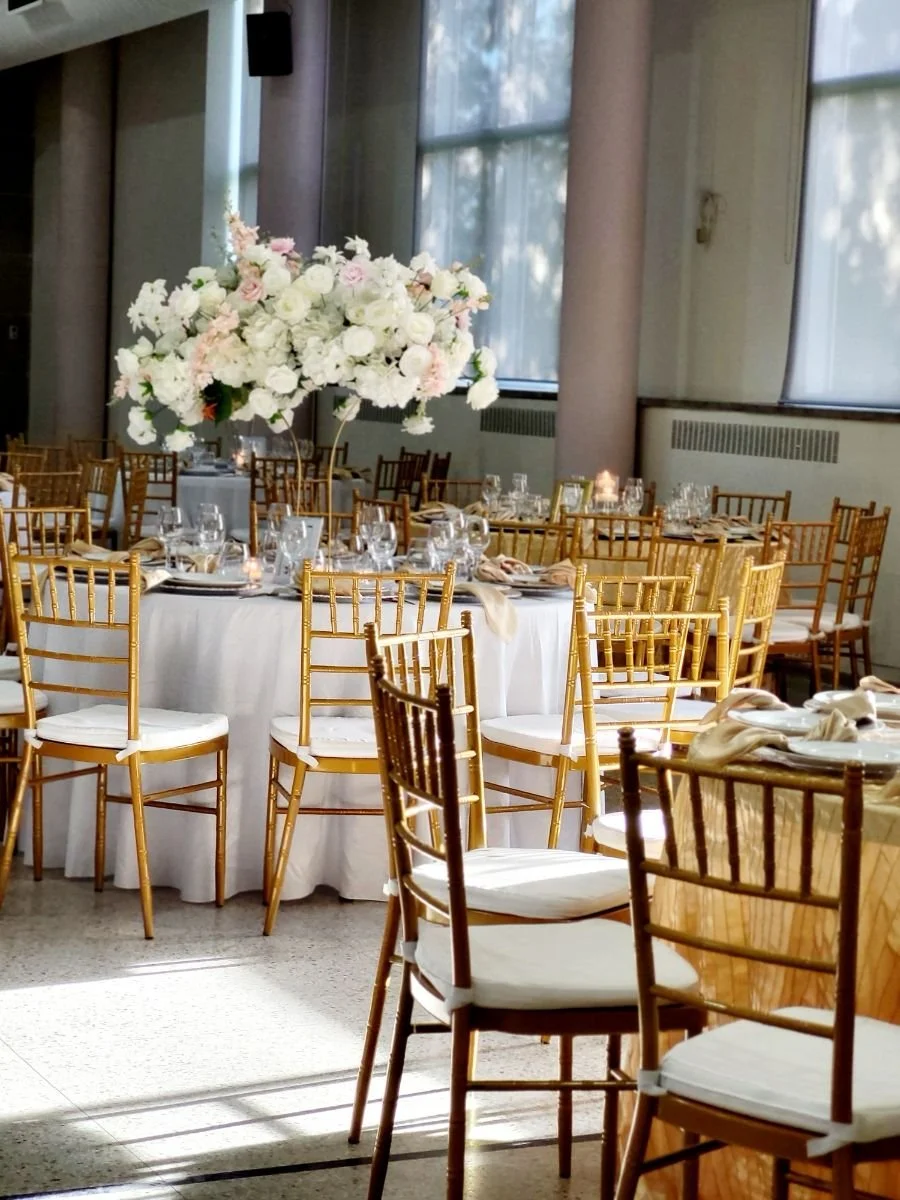 Elegant banquet hall decorated with a large floral centerpiece and round tables set with glassware and napkins, illuminated by natural light through large windows.