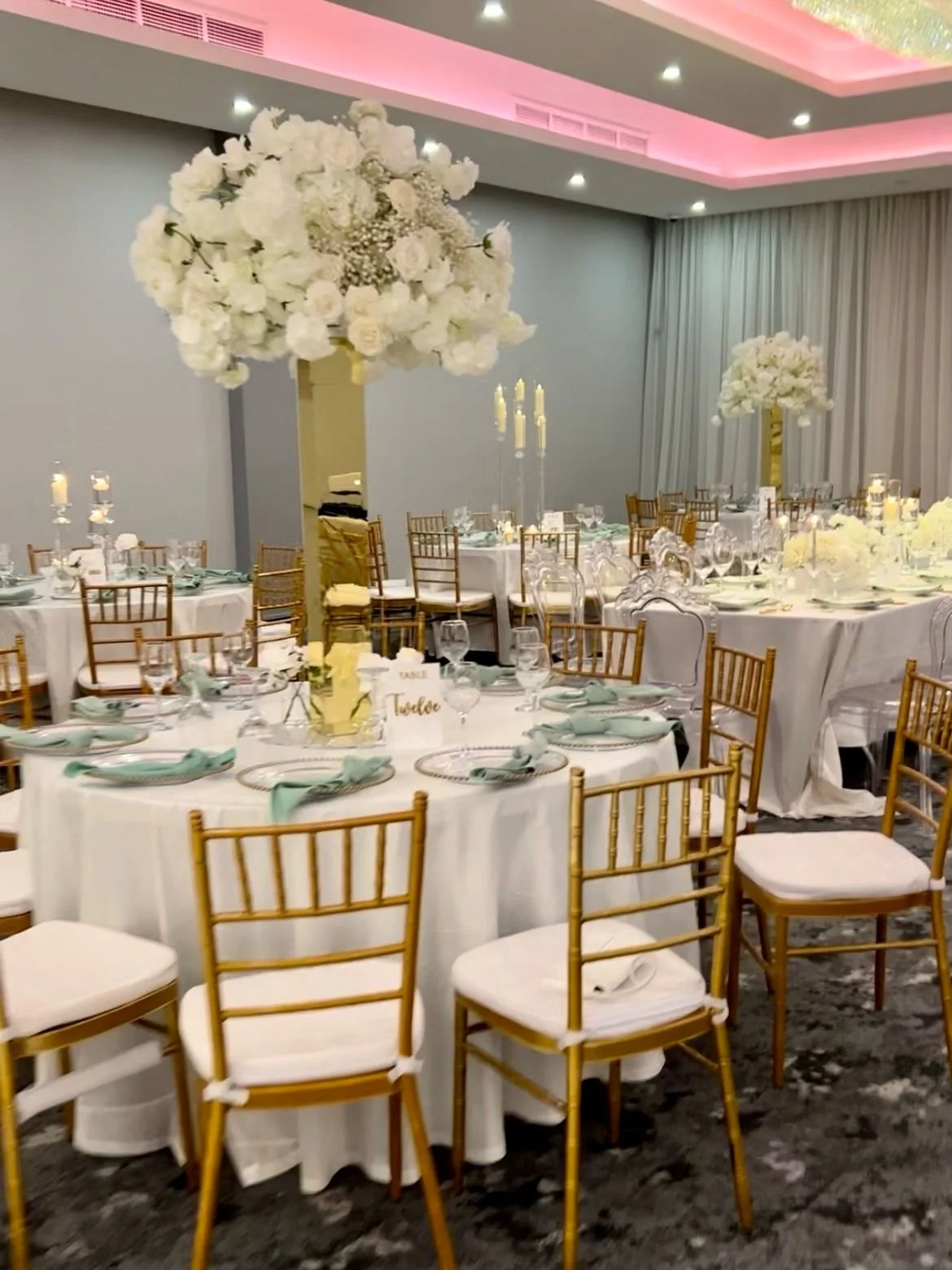 Elegant banquet hall decorated with white and gold accents, large floral centerpieces on tall vases, and round tables set with charger plates, glassware, and cloth napkins.