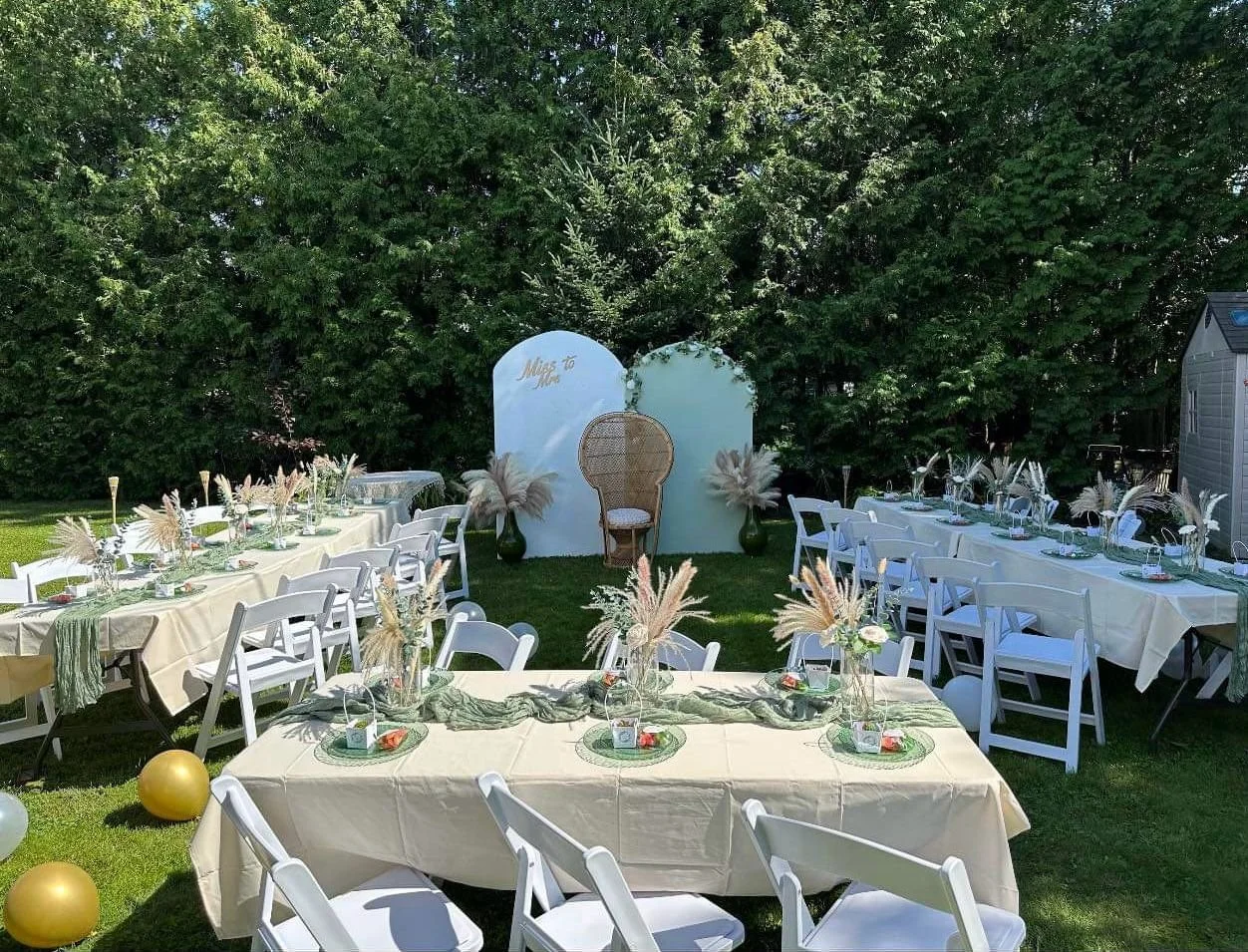 Outdoor wedding reception setup with long tables, white chairs, decorative centerpieces with dried flowers and pampas grass, on a green lawn with trees in the background, and a small backdrop with the words 'Miss to Mrs'.
