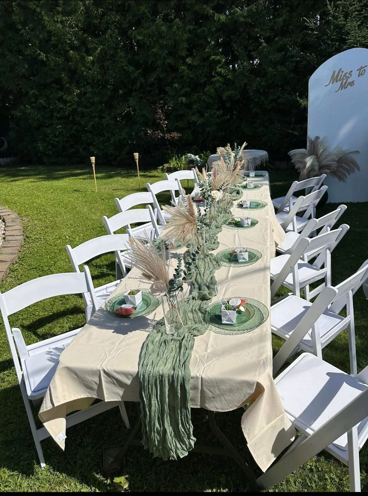Outdoor table set for a celebration with white chairs, a beige tablecloth, tall floral centerpieces, and decorative plates and favors, with a sign that reads 'Miss to Mrs' in the background.