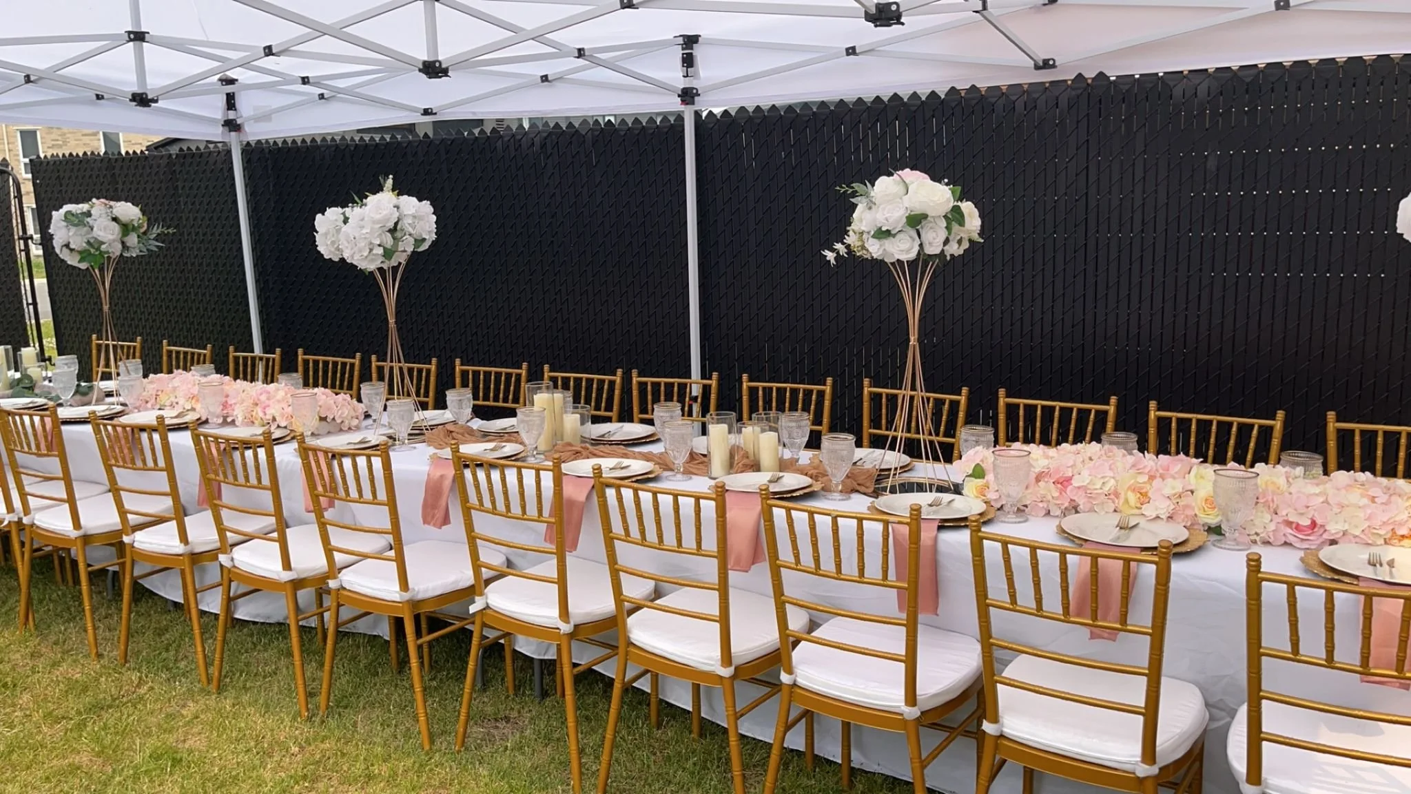 Long outdoor banquet table decorated with pink and white flowers, tall floral centerpieces, candles, pink napkins, and gold chairs under a white canopy.