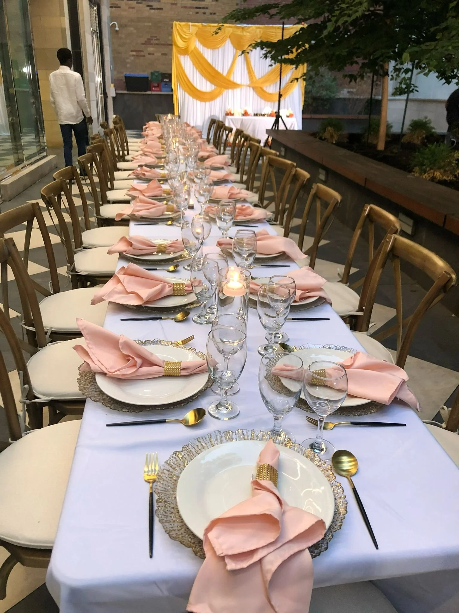 Long banquet table set for a celebration with pink napkins, gold utensils, clear glasses, and decorative plates, in a well-lit outdoor venue with yellow and white drapery at the back.