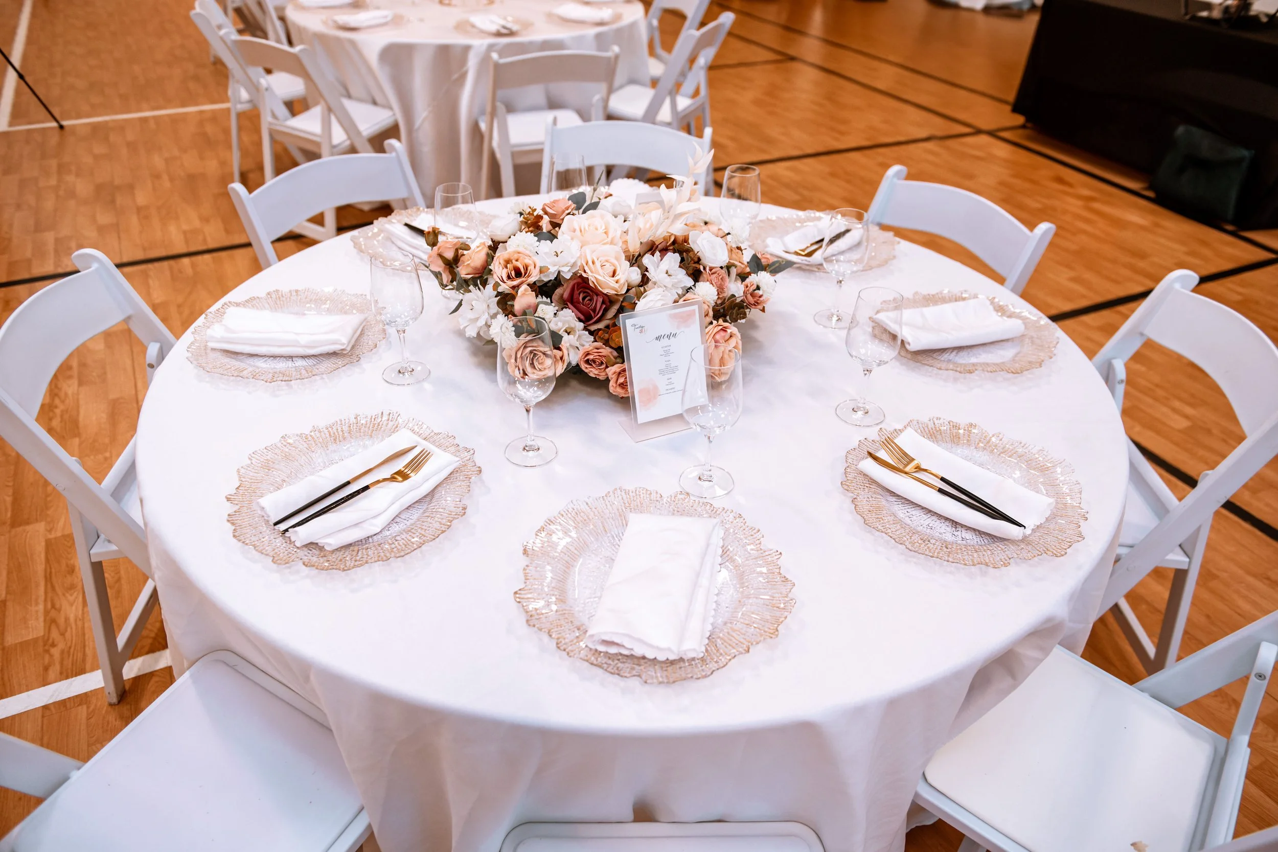 A round banquet table set for a formal event with a white tablecloth, a central floral arrangement of roses and other flowers, and place settings with white napkins, gold and black cutlery, and clear wine glasses. The chairs around the table are whit