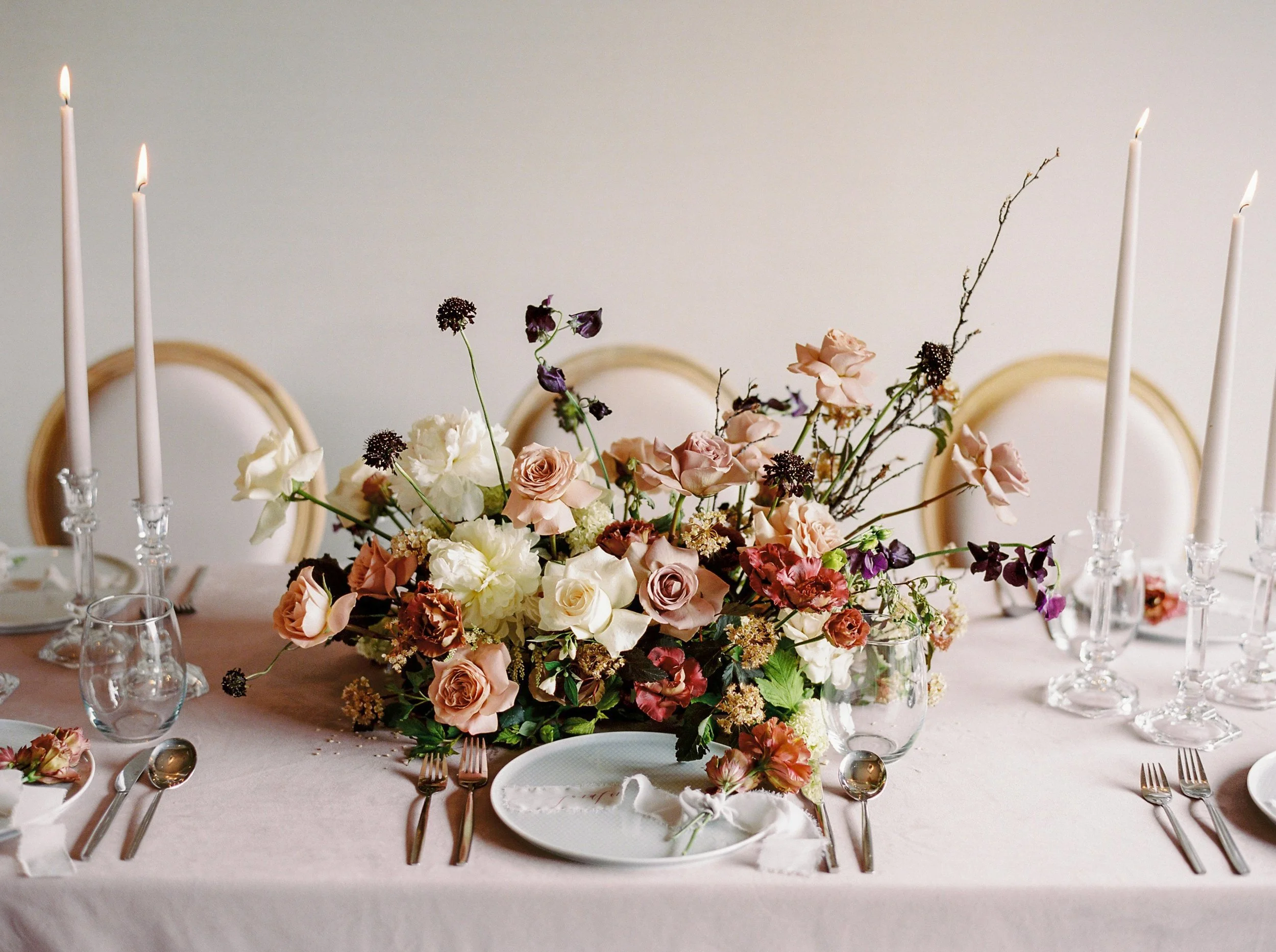 Elegant dining table with a large floral centerpiece featuring pink, white, and dark purple flowers, surrounded by glass candle holders with lit white candles, set with white plates, silverware, and wine glasses, on a light pink tablecloth.