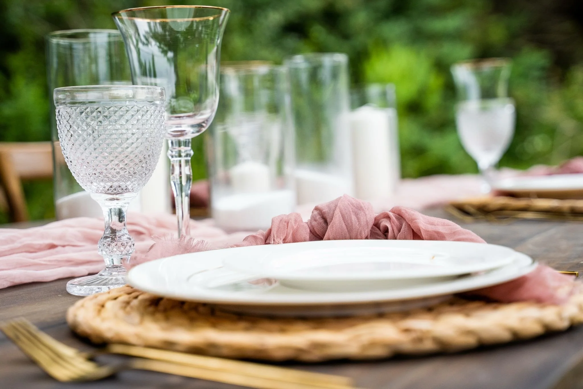 A set outdoor table with a woven placemat, white plates, pink cloth napkin, clear glassware including a cut glass wine glass and a taller glass, candles in white holders, and a gold fork, with a pink table runner and green foliage in the background.