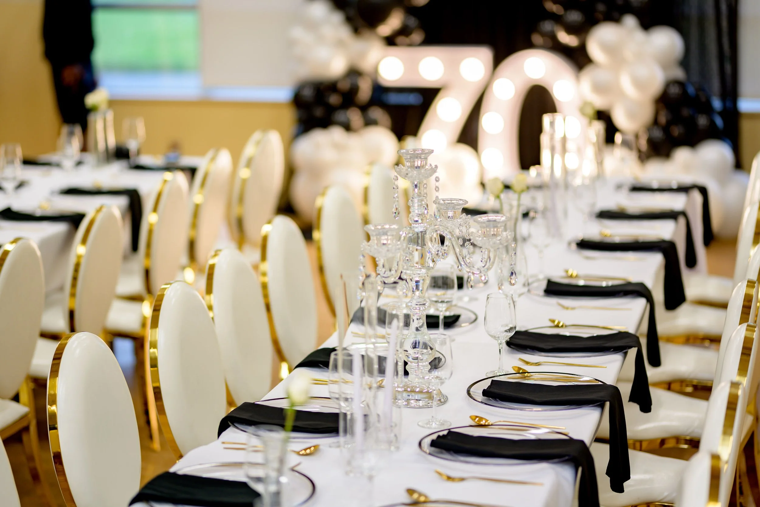 Long table set for a celebration with black and gold accents, gold cutlery, black napkins, and floral centerpieces, with a large illuminated '70' decoration in the background surrounded by black and white balloons.