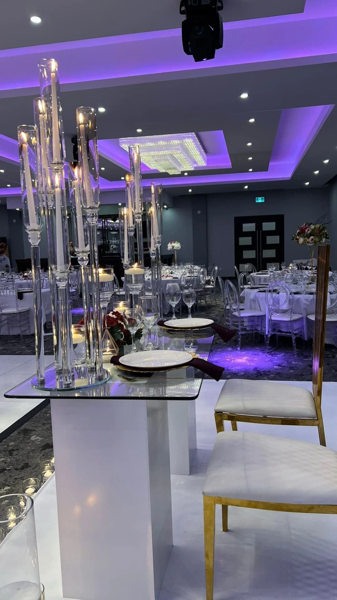 Elegant banquet hall decorated with tall glass pillar candles, floral arrangements, and set tables with white tablecloths, glasses, and plates, illuminated with purple lighting.