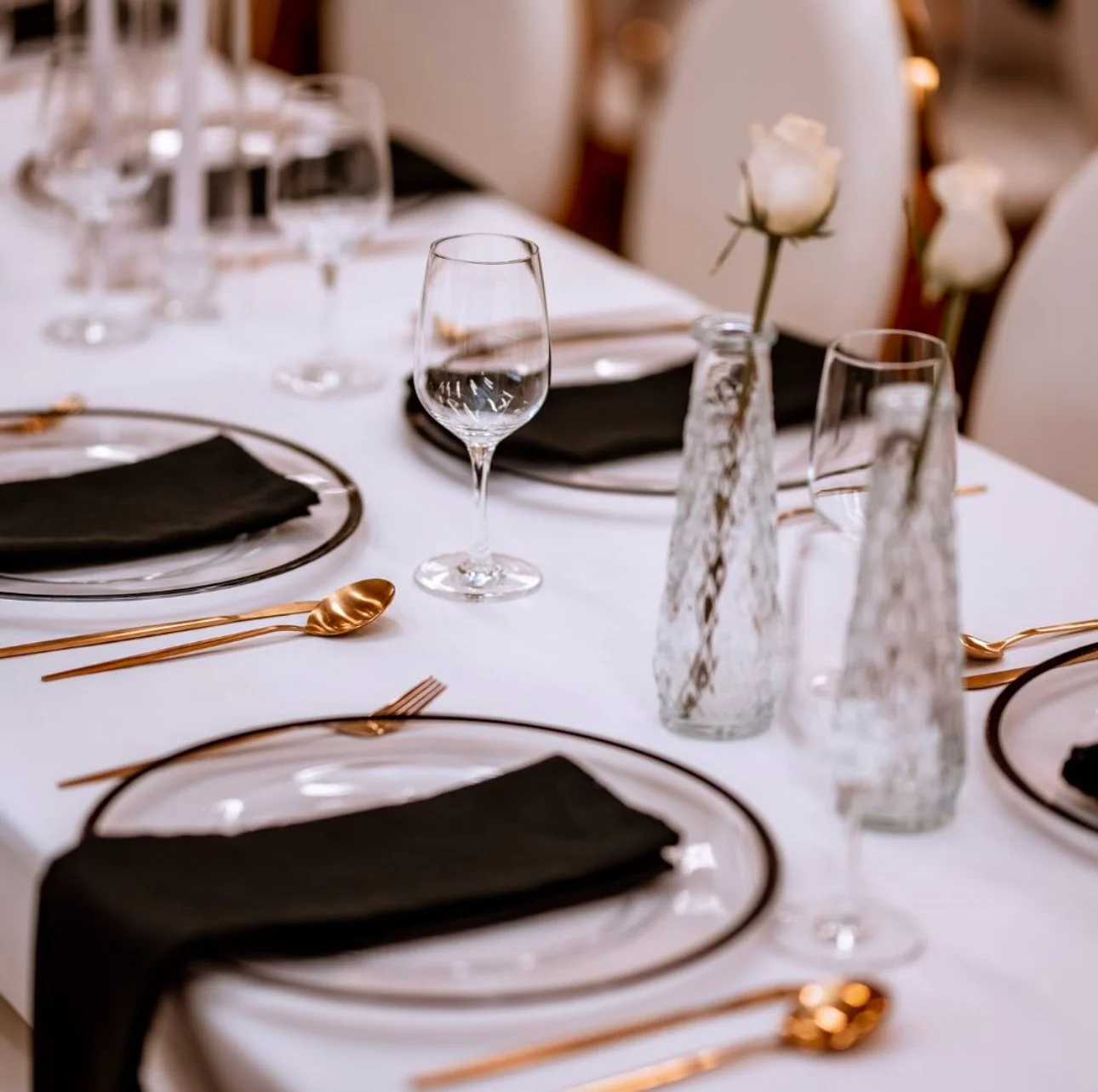 Elegant dining table set with black napkins, gold utensils, wine glasses, empty plates, and white roses in glass bottles.