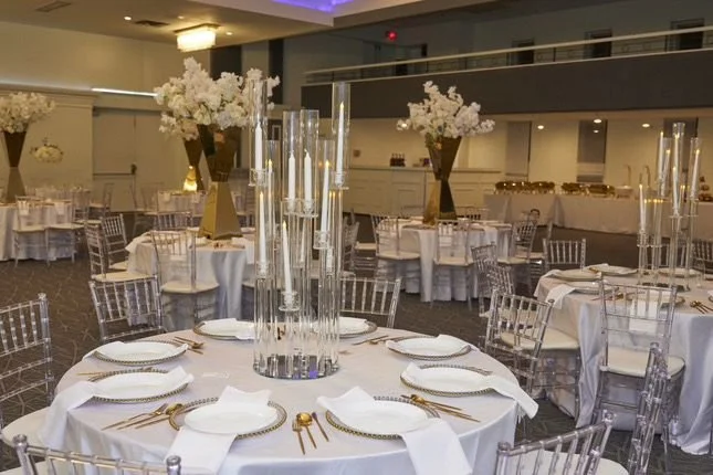 Elegant banquet hall with round tables set with white tablecloths, gold flatware, white napkins, tall glass candle holders, and large floral centerpieces with white flowers and gold vases.