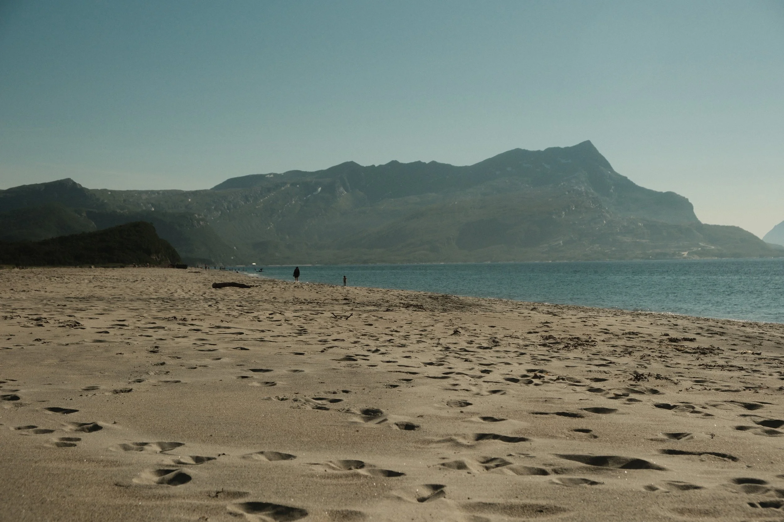 Sandsstrand med fjell i bakgrunnen, noen mennesker går langs stranden