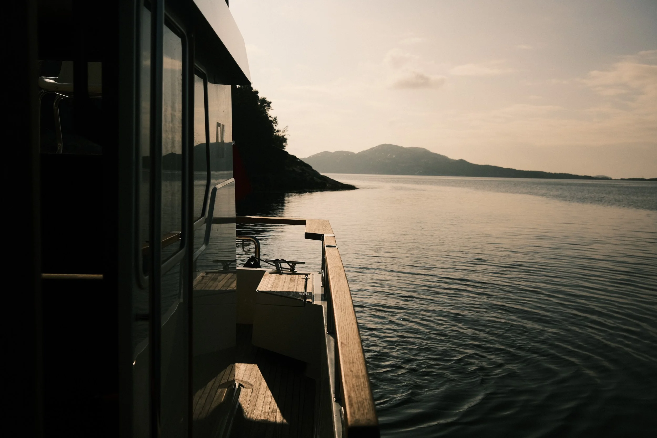Utsikt fra en båt med fjell og vann i bakgrunnen, solnedgang, og deler av båten i forgrunnen.