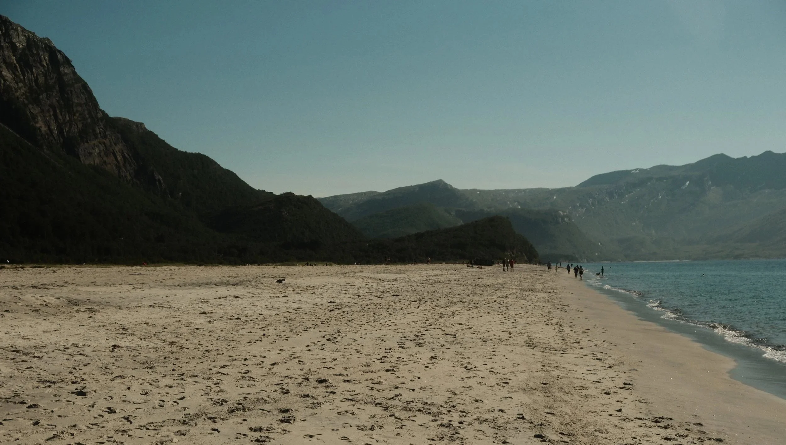 Bilde av en strand med sand og mennesker som går langs vannet, omgitt av fjell og en klar himmel.