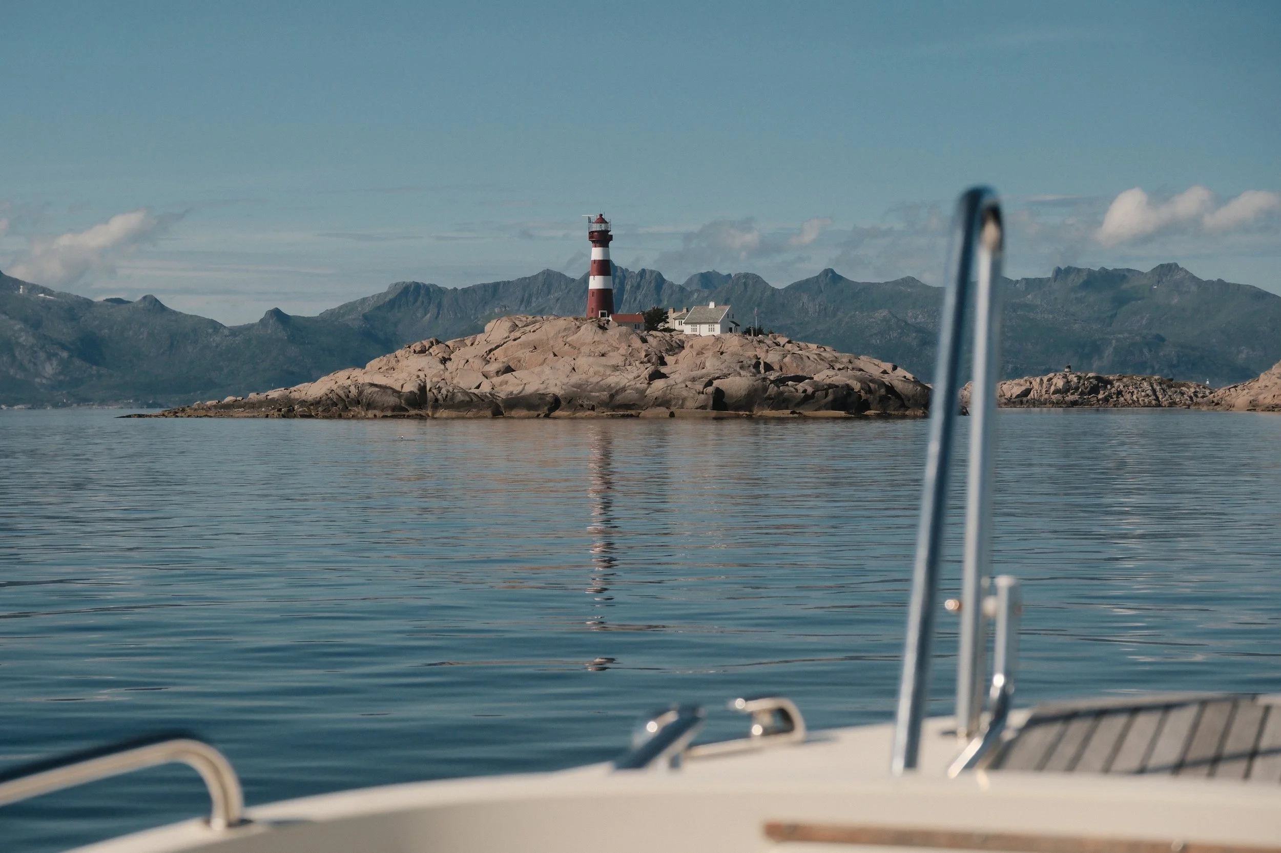 Utsikt over en sjø med en liten øy som har en rød og hvit fyrtårn, omgitt av fjell i bakgrunnen, sett fra en båts cockpit.