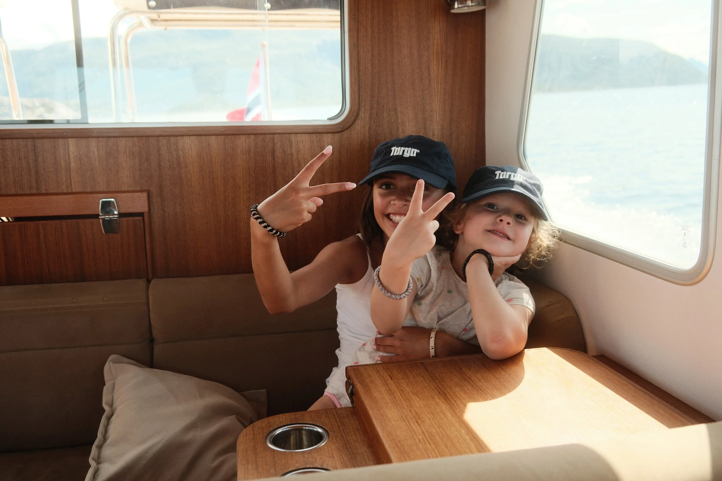 To unge jenter på en båt, smilende og gjør peace-tegn. De har på seg caps med logoen 'Targa'.