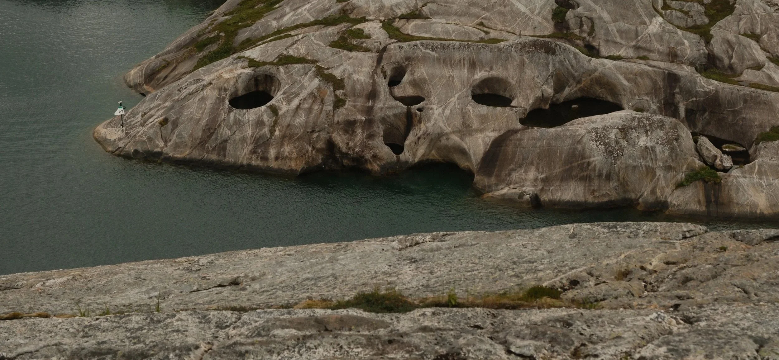 En stor stein med hull som ligner et ansikt, omgitt av vann, i en naturskjønn landskap.