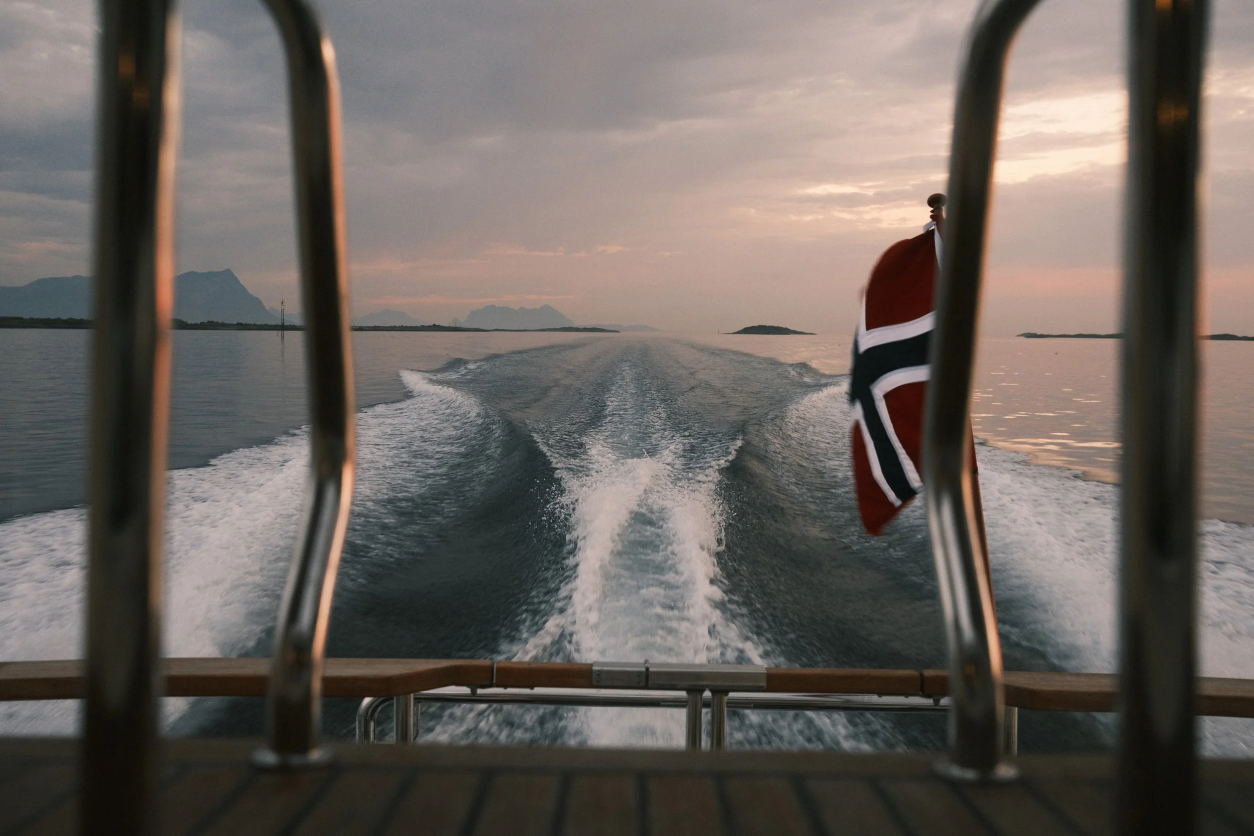 Utsikt fra en båt med vannsprut og sjølandskap, et norsk flagg flagrer i vinden, ved solnedgang.