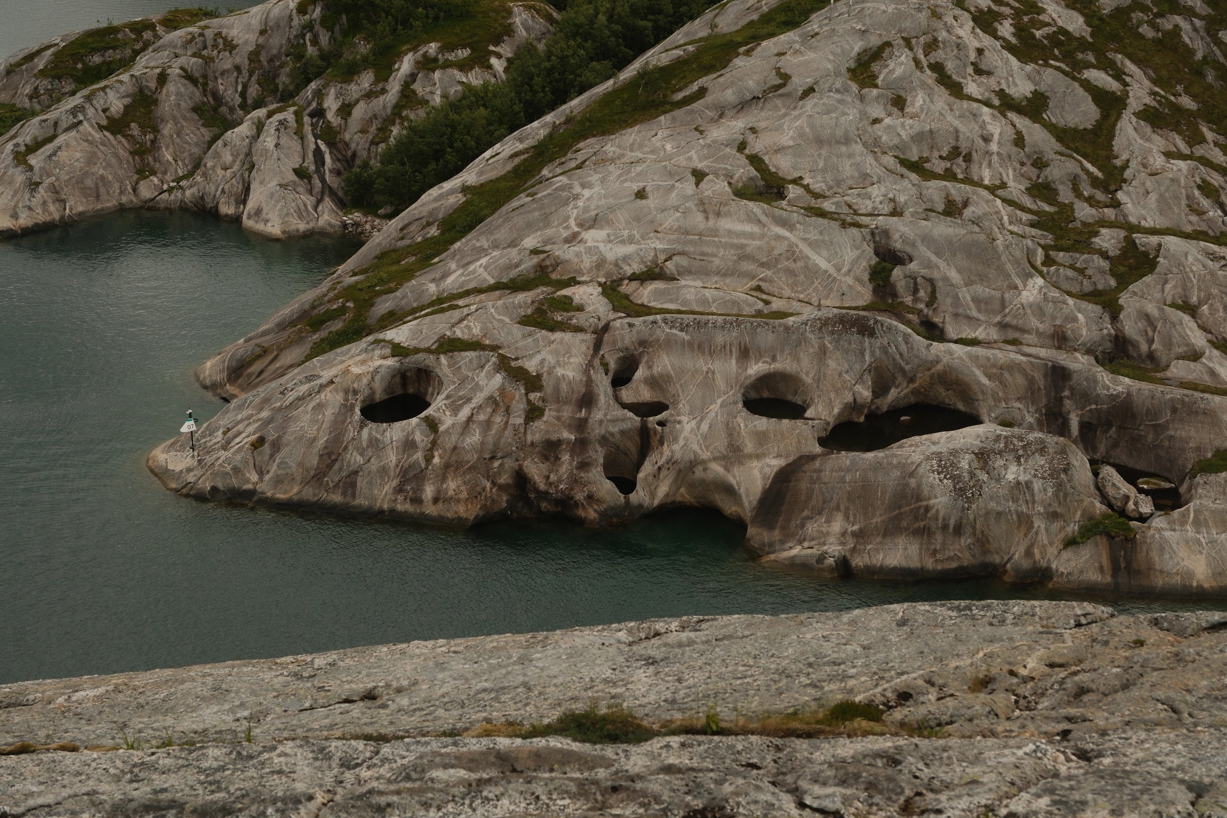 Stor bergformasjon med flere huler og hull, ligger ved vann med fjell i bakgrunnen.