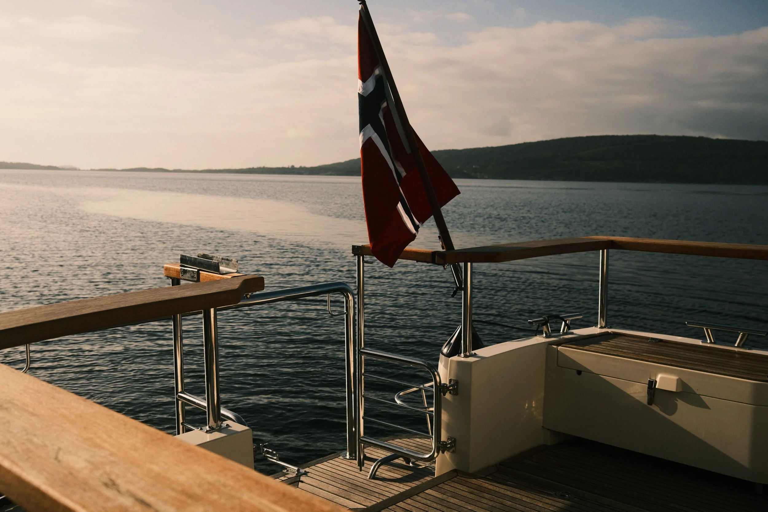 Utsikt fra en båt med et norsk flagg, utsikt over vann og fjell i bakgrunnen, solnedgang eller soloppgang.