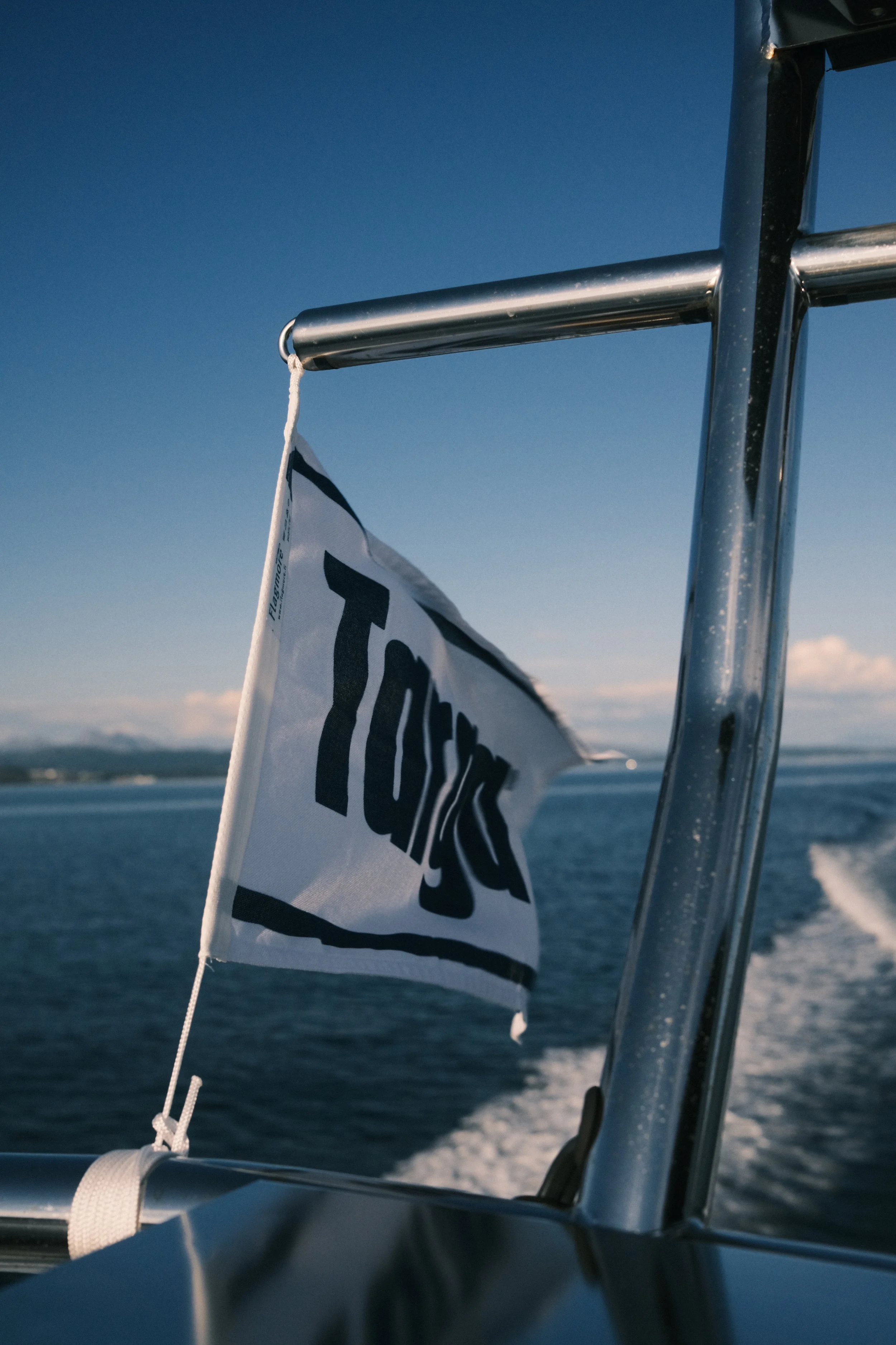 En liten flagg som sier 'Torsk' på en båt, med sjø og land i bakgrunnen.