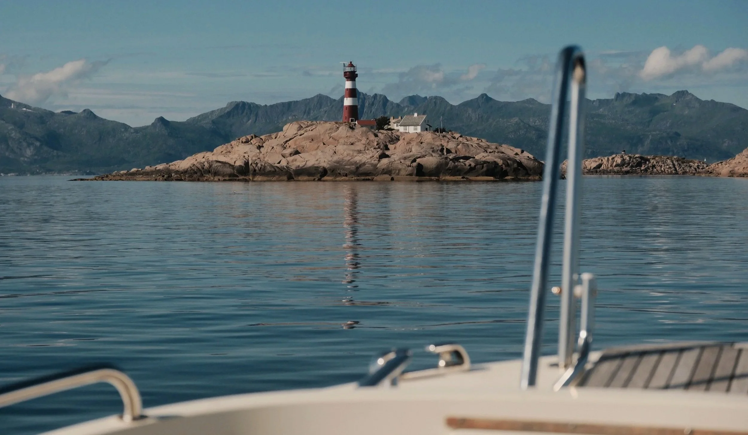 Utsikt fra en båt med en brygge i forgrunnen, et fyrtårn på en øy med steinete landskap, vann og fjell i bakgrunnen.