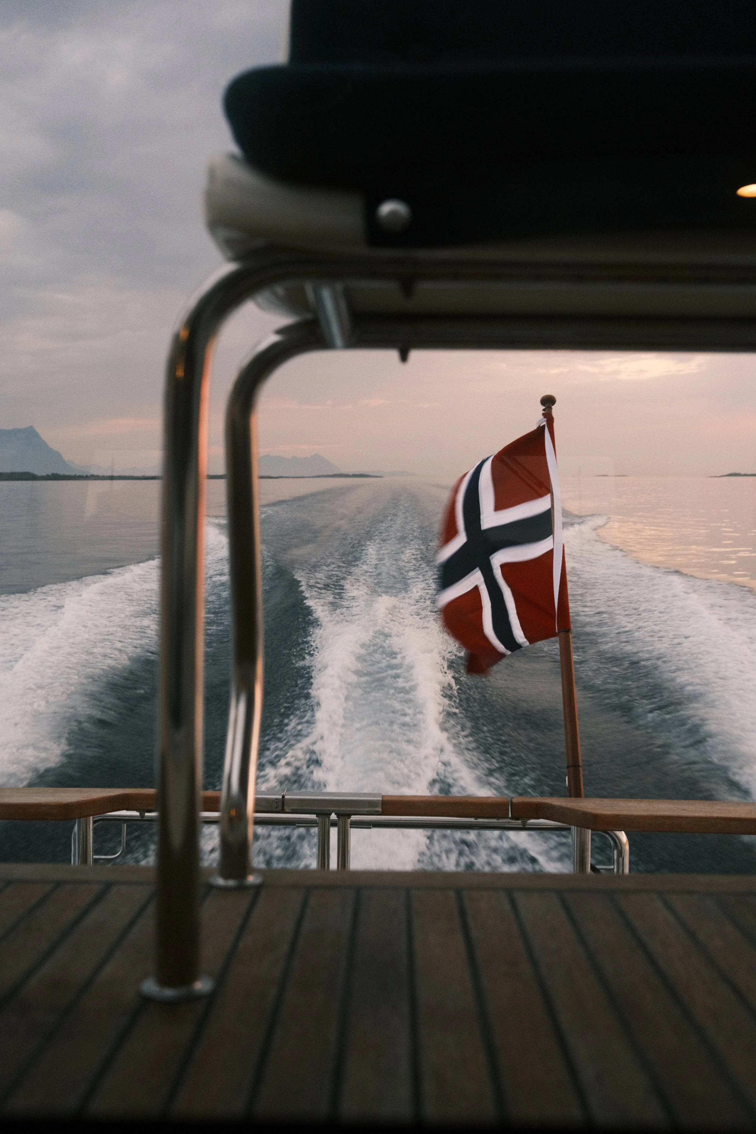 Utsikt fra båten med en norsk flagg, som flyr i vinden, på vannet under en solnedgang eller grå himmel.
