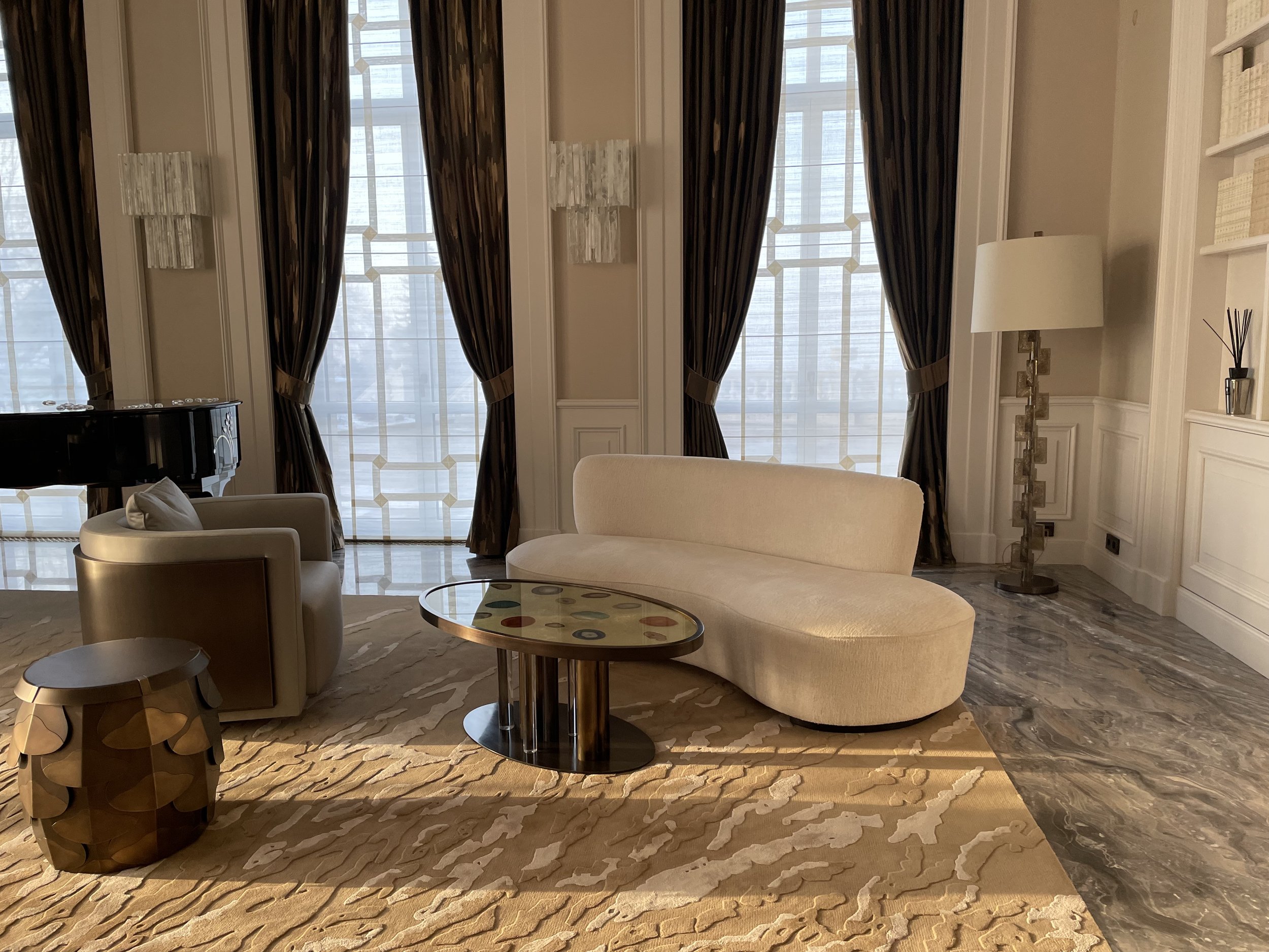 Un salon élégant avec des rideaux sombres, un canapé blanc incurvé, une grande lampe de sol, une table basse en verre et un piano à queue. Le sol est recouvert d'un tapis beige à motifs.