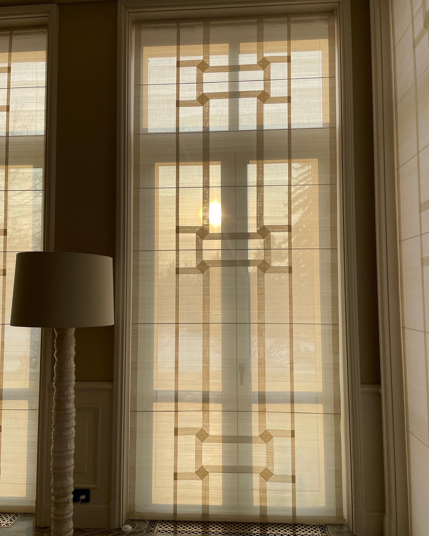 Panneau de vitres avec rideaux clairs et motifs géométriques, lampe à côté. Lumière naturelle entrant par la fenêtre.