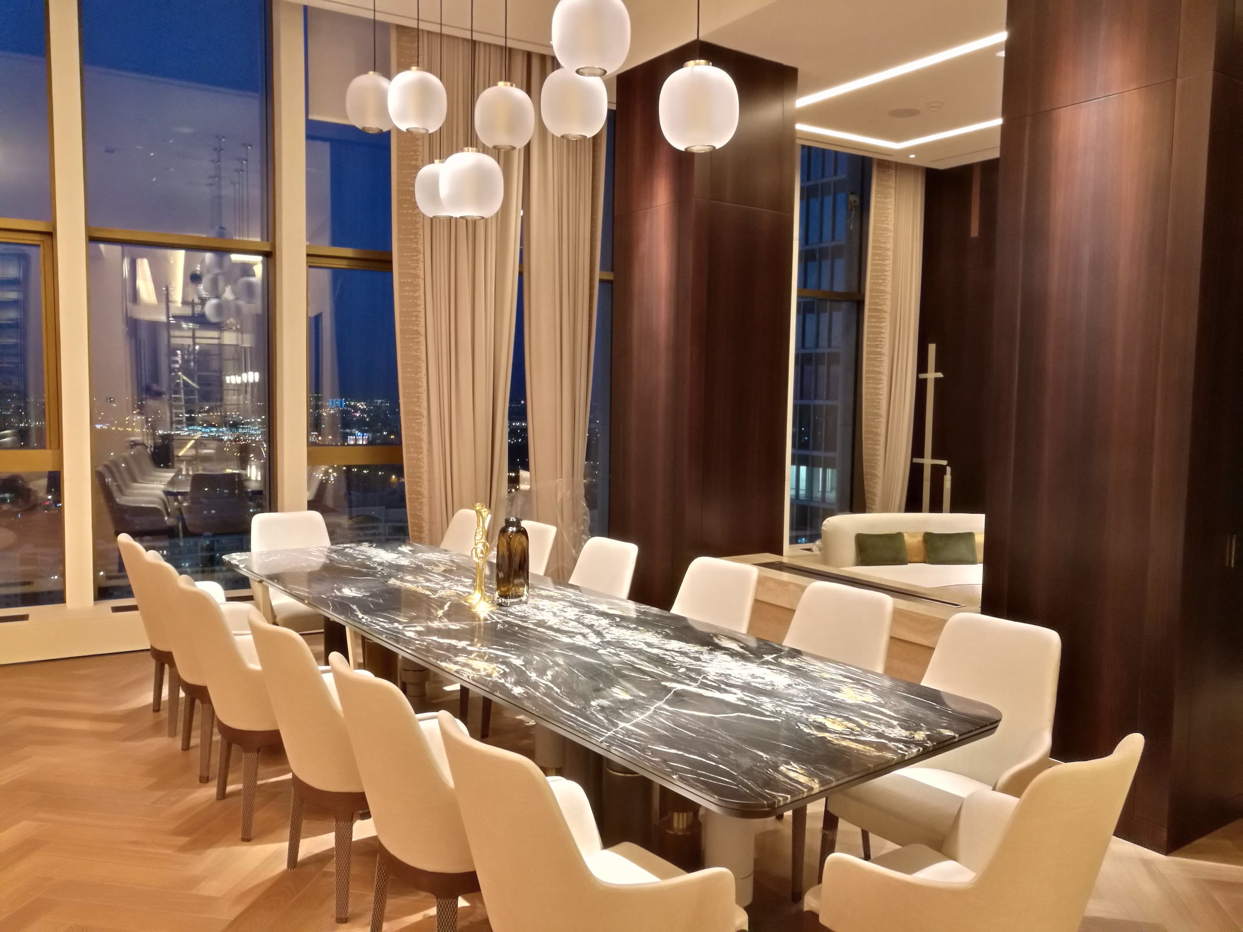 Salle à manger moderne avec une grande table en marbre noir entourée de chaises blanches, des lampes suspendues au plafond et des fenêtres offrant une vue sur la ville au crépuscule.