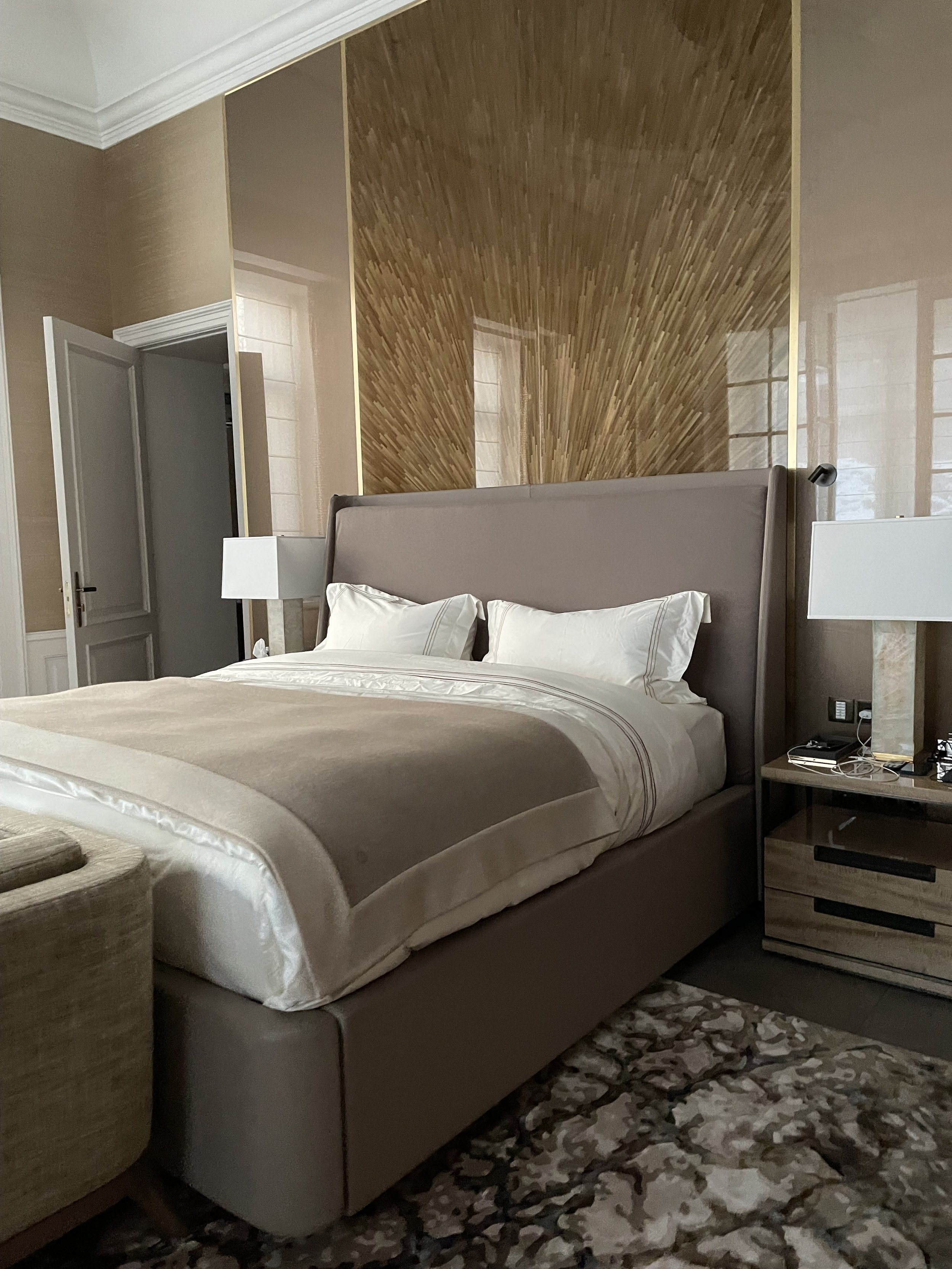 Chambre avec un grand lit luxueux à la tête rembourrée, murs décoratifs en bois, des lampes modernes de chaque côté, et un tapis à motif géométrique sur le sol. Il y a une table de nuit avec des objets personnels.