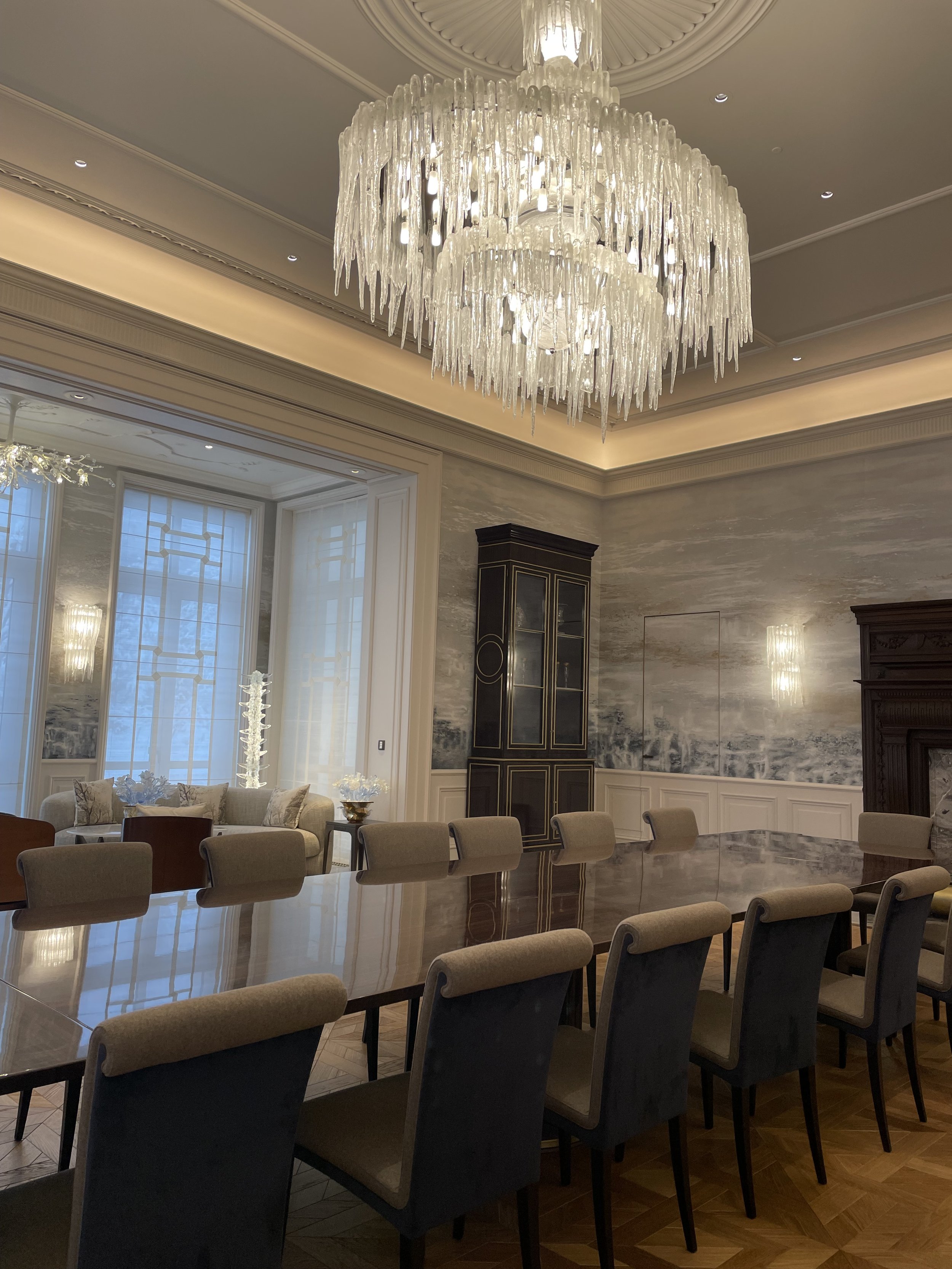 Salle à manger élégante avec un grand lustre en cristal, une longue table en bois entourée de chaises rembourrées, et une décoration murale sophistiquée.