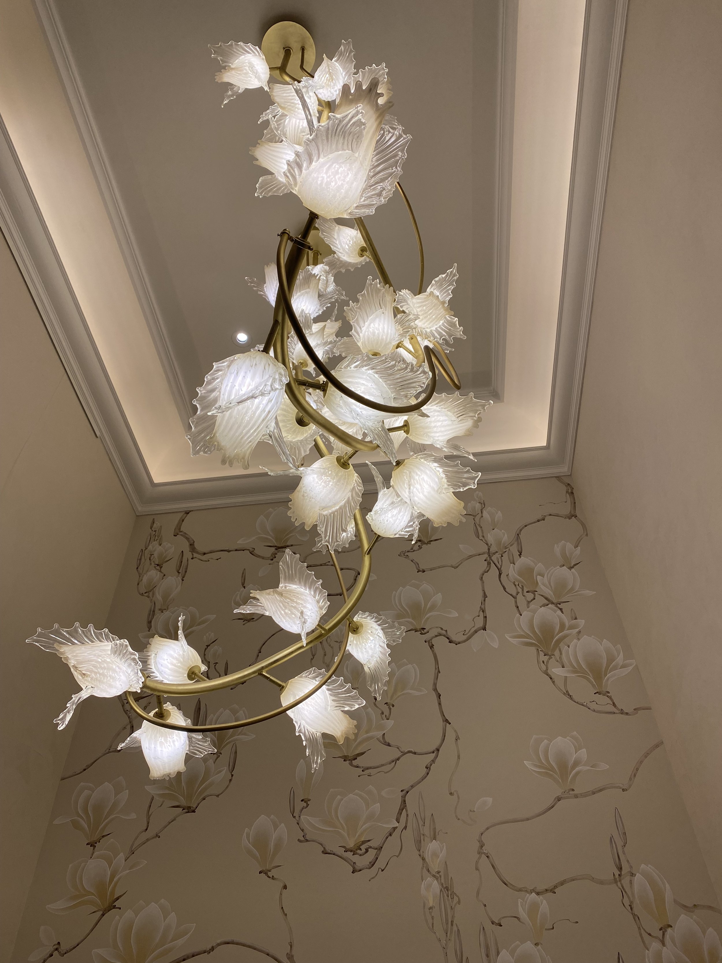 Un lustre floral avec des abat-jours en forme de fleurs blanches suspendu au plafond, sur un fond de papier peint avec des motifs floraux.