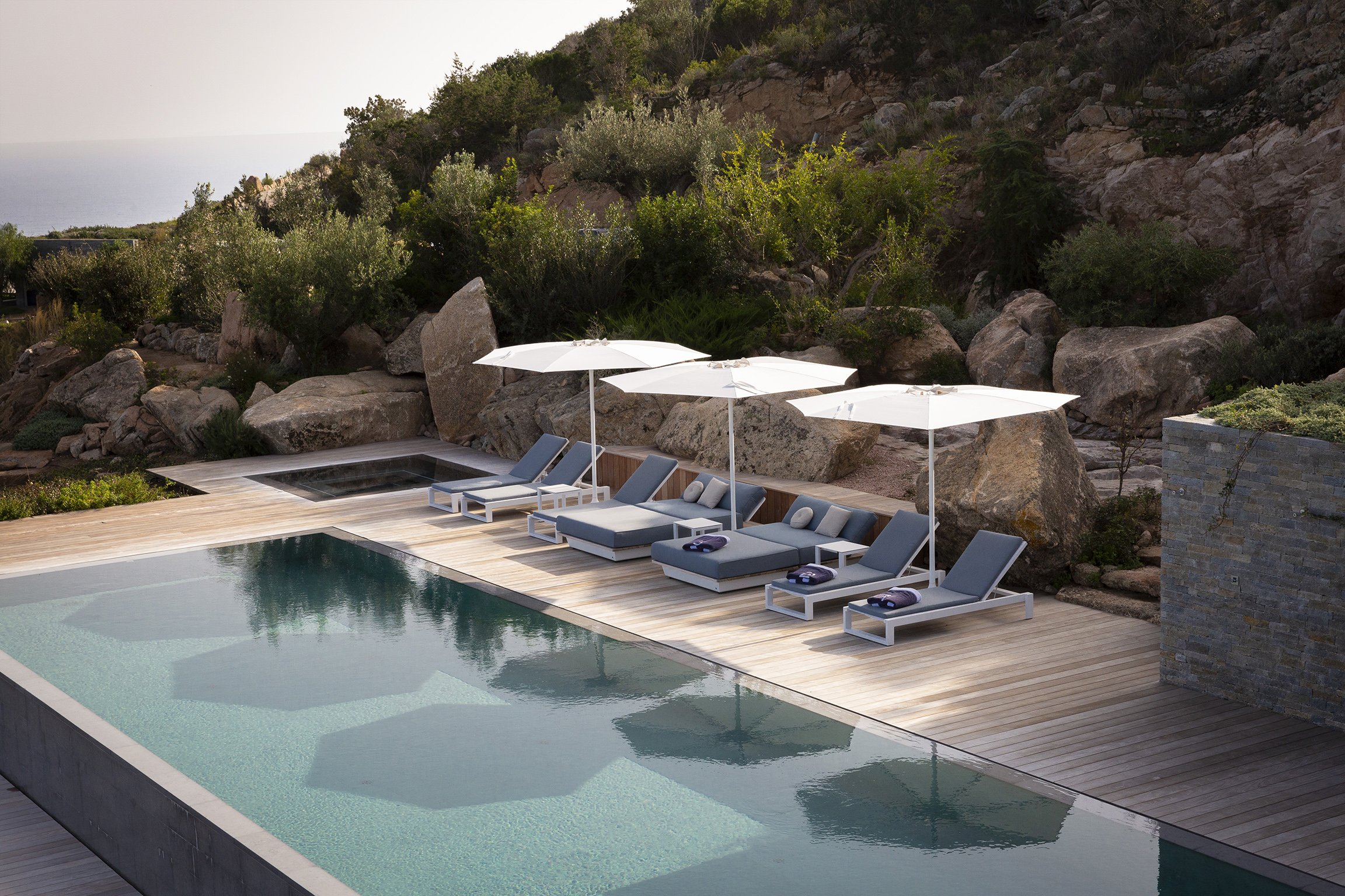 Piscine entourée de chaises longues avec parasols, sur une terrasse en bois, avec paysage rocheux et végétation environnante.