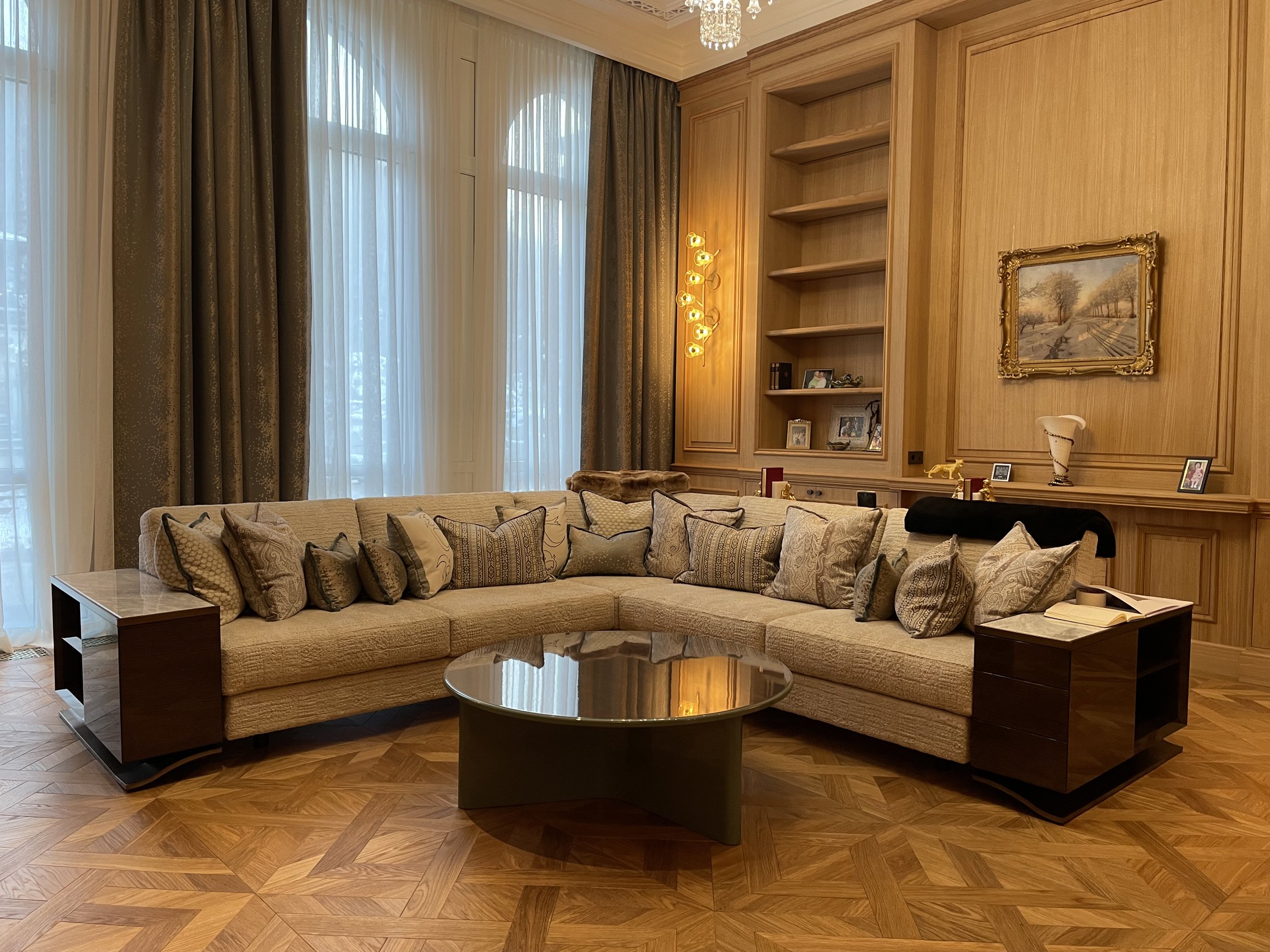 Un salon élégant avec un grand canapé beige orné de nombreux coussins, une table basse en verre, un parquet en bois, de grandes fenêtres avec rideaux, et une bibliothèque avec des étagères, décorée de cadres et d'objets d'art.
